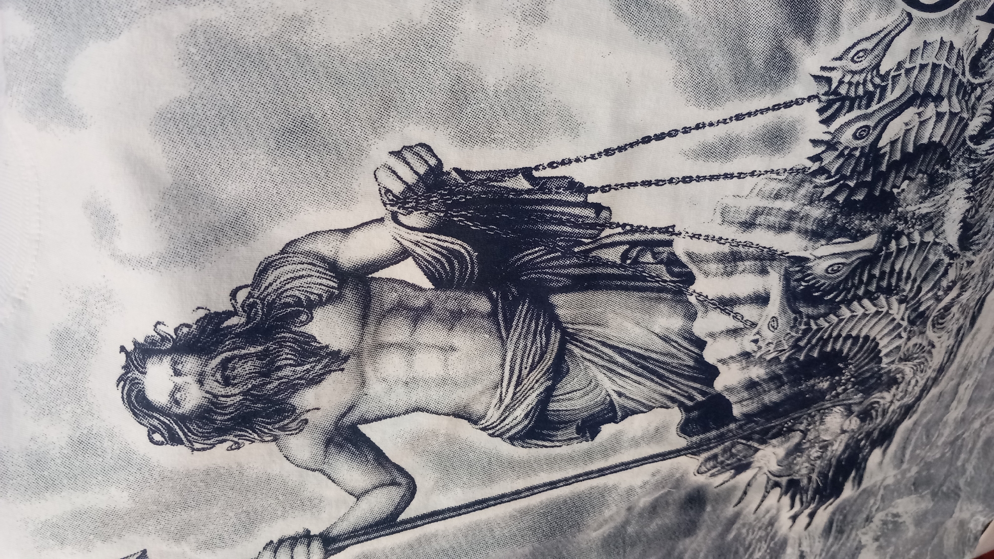 The image is a black and white graphic print of a muscular, bearded man with long, wavy hair, standing in rough water. He holds a trident in his left hand and a draped cloth with chains in his right. He is positioned on a chariot-like structure being pulled by several seahorse-like creatures, also rendered in black and white. The background is a mottled grey, suggesting a textured fabric, likely a t-shirt. The graphic depicts a mythological figure, possibly Poseidon or Neptune, in a dynamic maritime scene. The image does not contain any identifiable landmarks or features that would place it in the city of Kos, Greece.