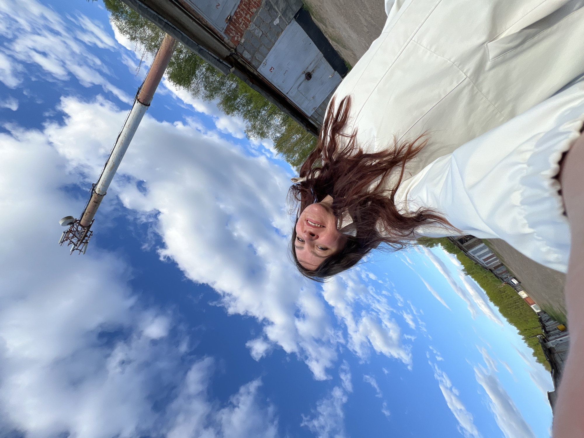 The image shows a woman taking a selfie in Noviy, Russia. She is wearing a white coat. Behind her is a bright blue sky with fluffy white clouds, and a cell tower is visible on the left side of the frame. In the background, there are rows of garages with brick and metal doors. A line of trees can be seen above the garages, suggesting an outdoor environment.