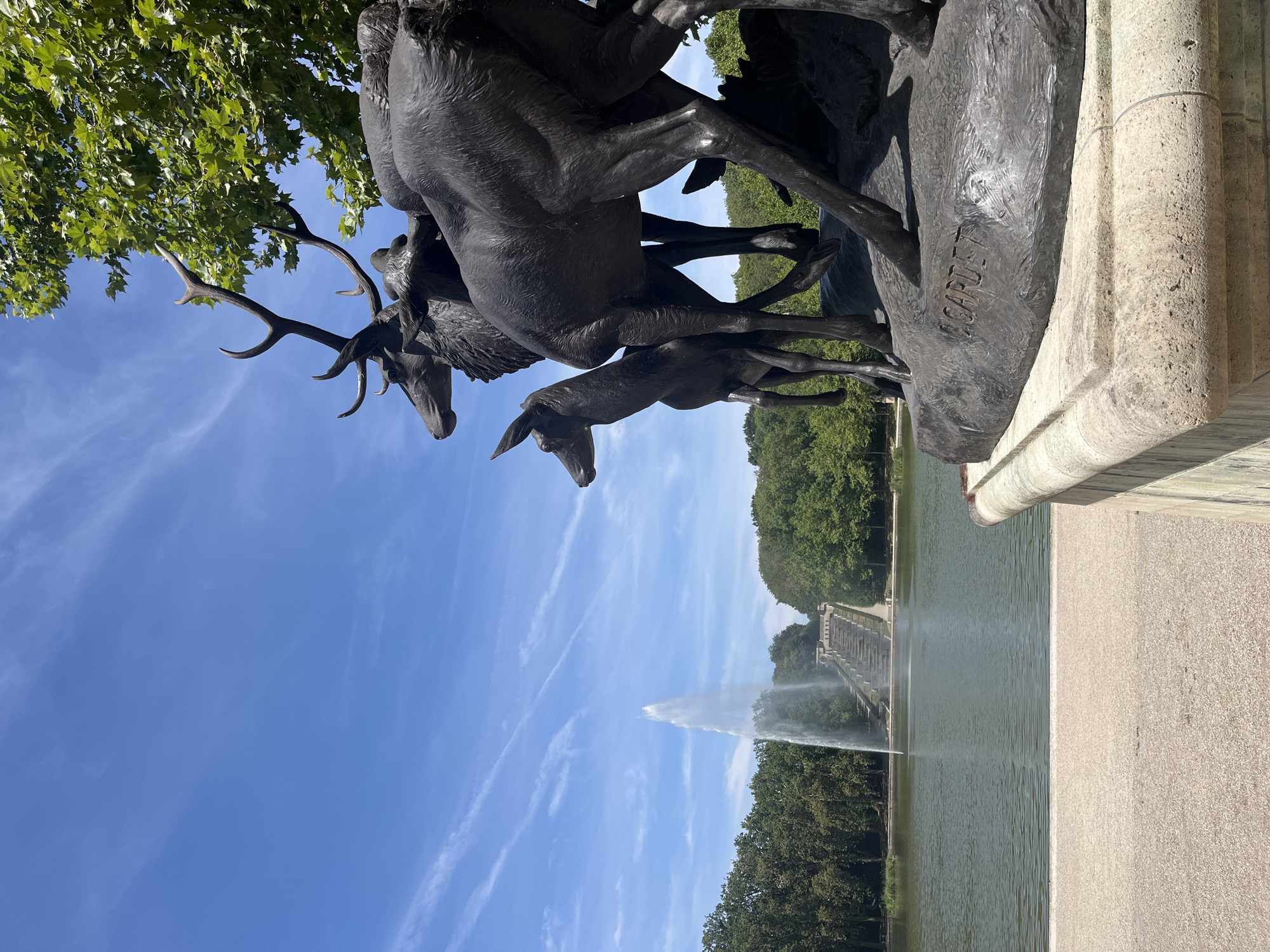 A bronze sculpture of several deer stands in the foreground on the right side of the frame, on a stone pedestal. In the midground, a large body of water reflects the sky, with a tall fountain spraying water upwards in the center. Beyond the water, a line of trees forms a dense green backdrop. Further in the distance, a grand staircase with multiple tiers leads up towards the horizon. The sky is a clear blue with scattered wispy white clouds. The overall scene depicts a serene park or garden setting. This is the Fountain of the Deer in Parc des Sceaux, located in Antony, France.