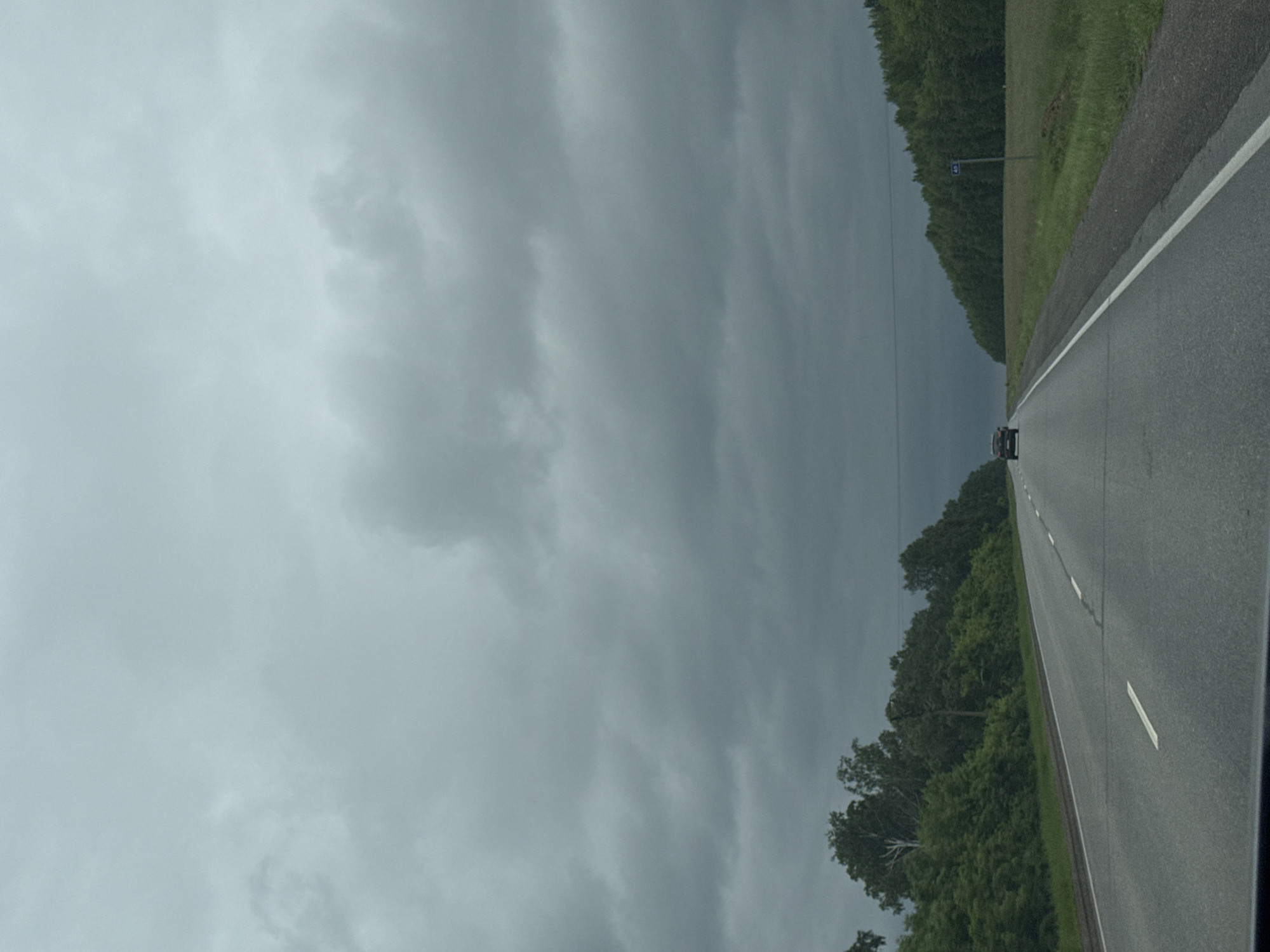 The image depicts a view from a vehicle traveling on a two-lane highway in Старошареево, Russia. The sky is heavily overcast with dark, heavy clouds, suggesting an impending storm or overcast weather. The road stretches forward, with a white line marking the center and a white line on the right edge. On both sides of the road, there are dense green trees forming a thick forest, with a grassy field visible on the right side. A single dark-colored car is visible in the distance, traveling in the same direction as the viewer. A blue road sign with the number "40" is present on the right side of the road. The overall atmosphere is somber due to the weather conditions.