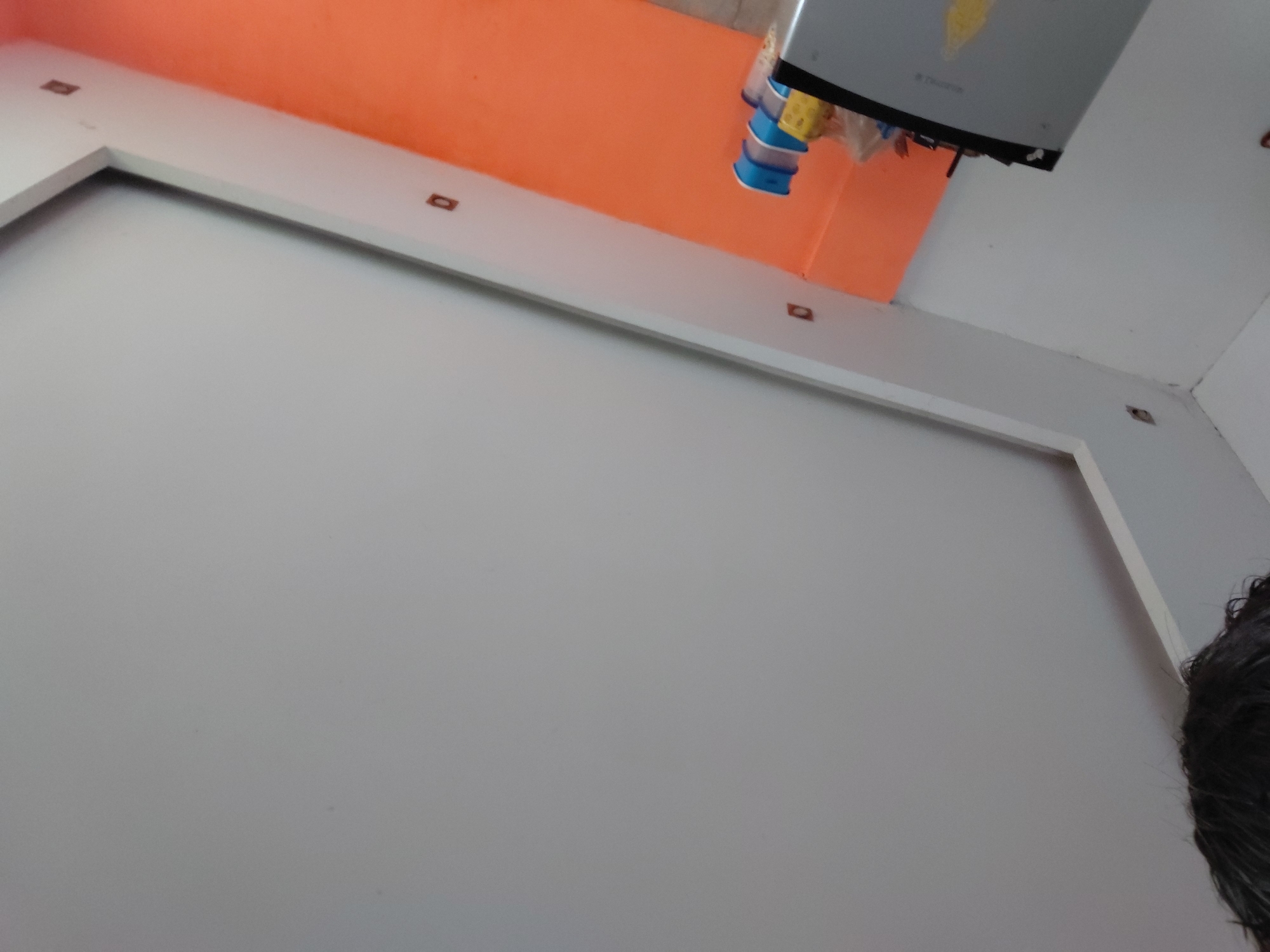 This image provides an upward-angled view of an interior room, likely in Santa Ana, Venezuela. The primary subject is a large, plain white wall that takes up the majority of the frame, bordered by a distinct white architectural trim or frame. Above this white section, an orange wall or ceiling is visible, creating a contrasting background.
Several small, rectangular brown objects are embedded within the white trim, resembling recessed lights or perhaps electrical outlets. These are distributed along the trim's horizontal and vertical segments.
Hanging from the upper part of the ceiling or wall is a grey, rectangular appliance, possibly a small water heater or similar utility device. This appliance features blue and yellow components attached to its underside. Faint, unreadable text, possibly a brand name with visible letters that could be "TRUTON" or similar, is present on its grey surface.
In the bottom right corner, the top of a person's head with dark, curly hair is partially visible, suggesting the perspective of someone looking upwards. No specific activity or event is taking place; the scene is static, capturing details of the room's structure and contents. The lighting is even, typical of an indoor setting with diffused light.