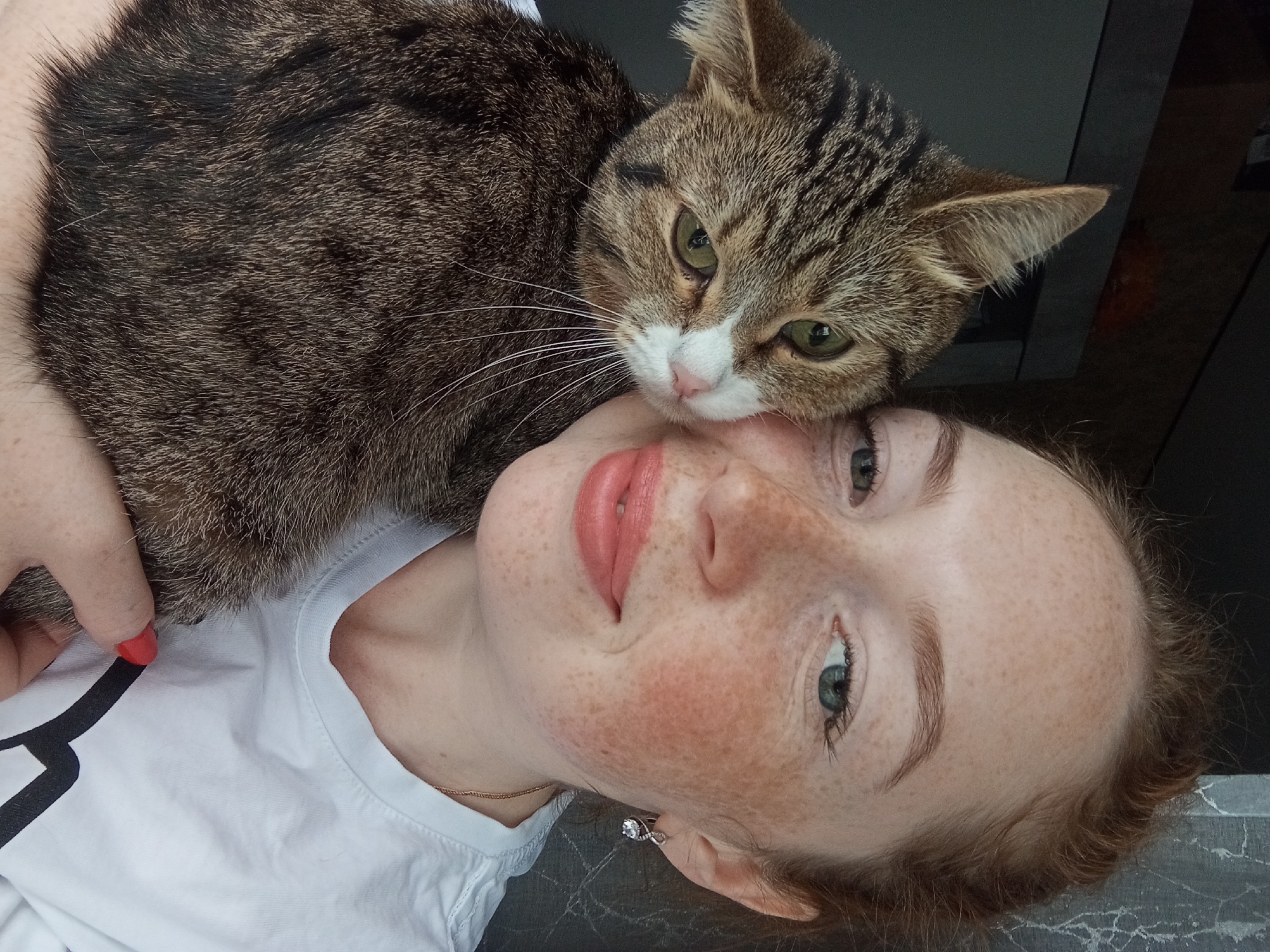 A woman and a cat are pictured in a close-up, overhead shot. The woman is lying on her back, wearing a white t-shirt with black chevron details on the shoulder. She has freckles, light eyebrows, and fair skin. The cat is resting on top of her, near her face. The cat is a brown tabby with a white muzzle and chest, and its green eyes are visible. The background appears to be a dark, reflective surface. The location is Yurgamysh, Russia.