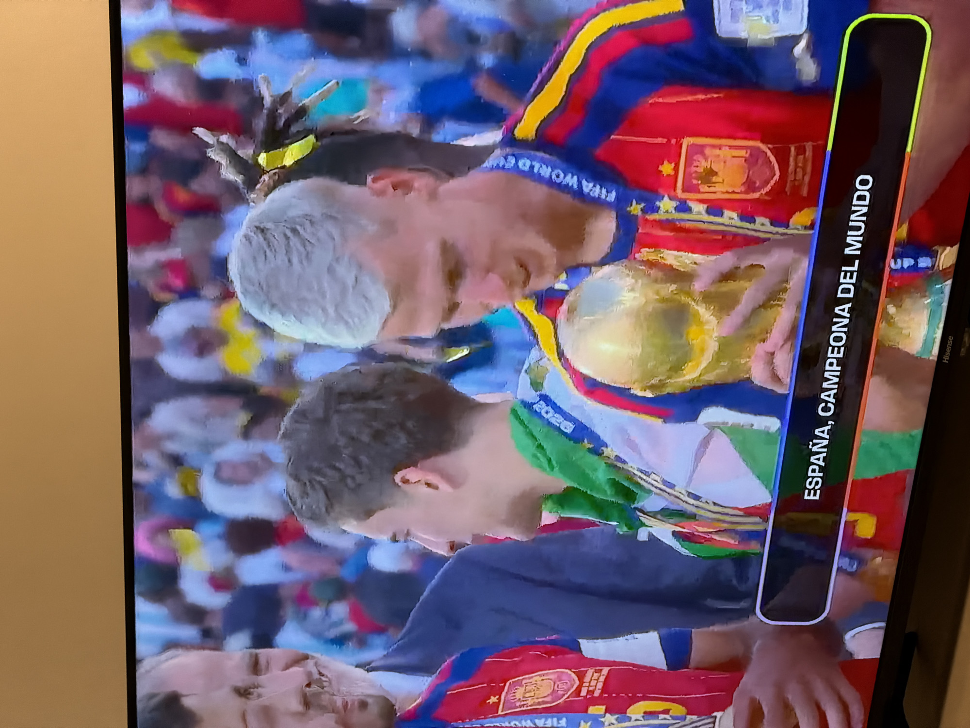 The image captures a screen displaying a television broadcast, likely a sports event. In the foreground, two men are visible, one with short, light-colored hair and the other with darker, curly hair. The man with light hair is wearing a red and blue striped jersey with yellow accents and a medal around his neck. He is holding a golden trophy. The man with darker hair is wearing a green scarf around his neck over a white and green jersey, also adorned with medals. Behind them, a crowd of people in various colored clothing is visible, suggesting a celebratory atmosphere. A banner at the bottom of the screen displays text that reads "ESPAÑA, CAMPEONA DEL MUNDO." The television set itself rests on a surface, with a black bezel and a visible brand name "Hisense" at the bottom. The background wall appears to be a pale yellow. While the image is of a broadcast showing a team identified as "España" (Spain), there is no visual information within the image that specifies a location within the city of Finestrat, Spain.