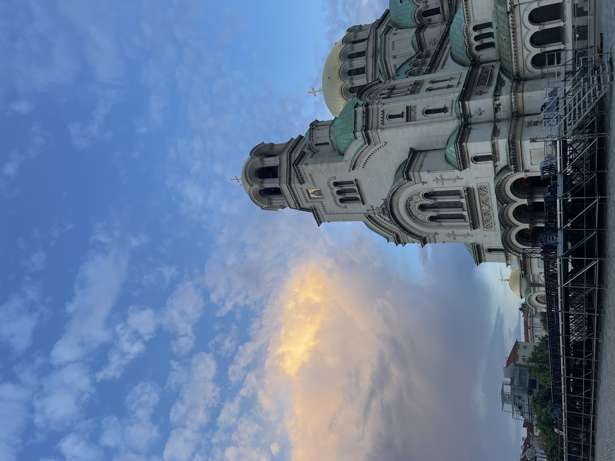 A towering Eastern Orthodox cathedral dominates the right side of the frame, captured under a dramatic sky, likely at sunset or sunrise. The cathedral, built from light-colored stone, features multiple prominent domes; some are gilded gold, while others are a distinctive teal-green. Each dome is topped with an Orthodox cross. The structure showcases intricate architectural details, including arched windows, decorative reliefs, and a tall bell tower with an icon of a saint visible on its facade.
The sky above transitions from a clear blue with scattered white, wispy clouds at the top, to a more complex and colorful cloudscape lower down. A striking cloud formation, centrally positioned in the mid-ground, is illuminated by a warm, golden light, contrasting with the surrounding greyer, diffused clouds.
On the far right, a large metal scaffolding or temporary structure stands, partially obscuring the lower sections of the cathedral. In the background, beyond the scaffolding, glimpses of smaller buildings and green trees are visible at street level. No people are discernible, and no specific event or activity is clearly depicted. No legible text is visible on the building or in the scene. The scene evokes a sense of grandeur and historical significance. The location context indicates this is the Alexander Nevsky Cathedral in Sofia, Bulgaria.