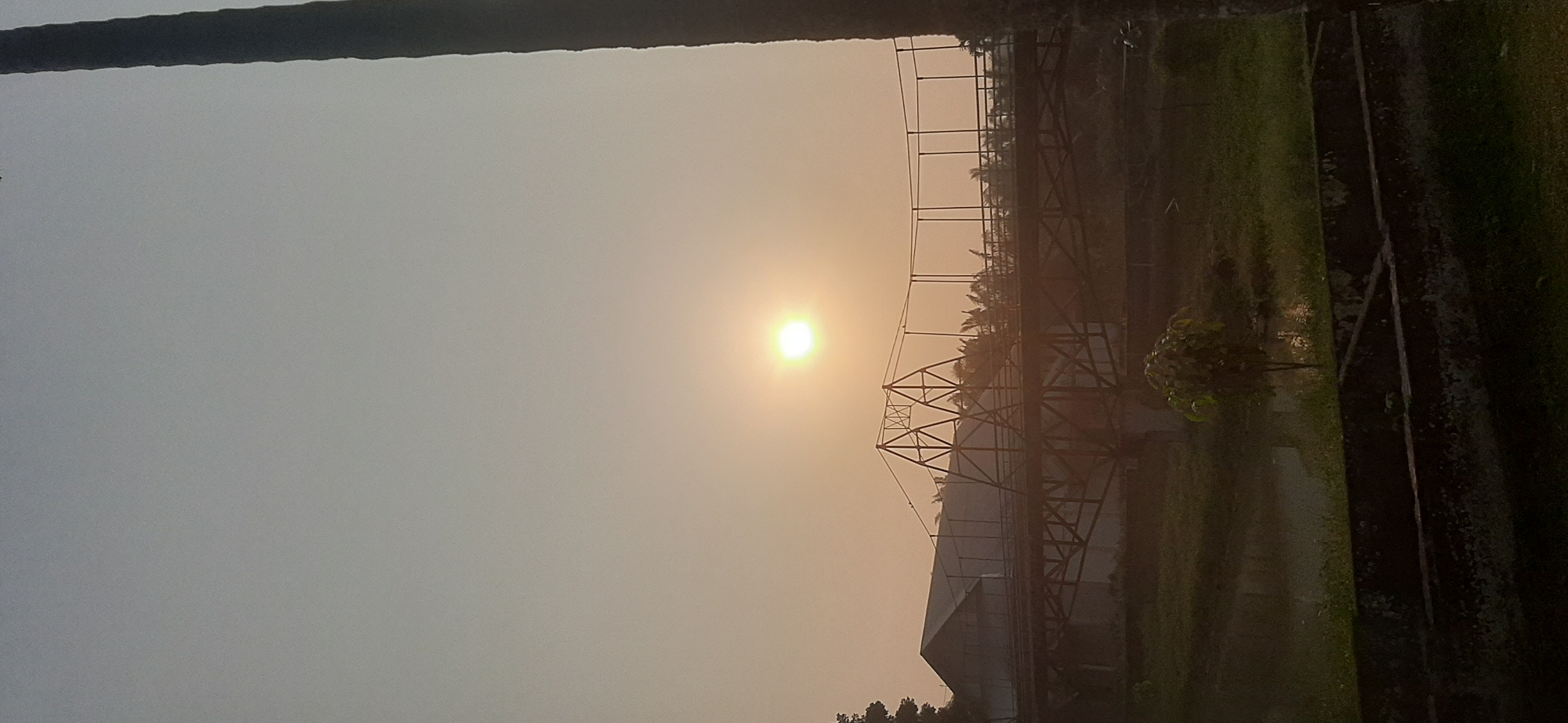 The image depicts a serene outdoor scene, likely Jambi, Indonesia, during a hazy sunrise or sunset. The sun, a bright orb, hangs in the pale orange sky, casting a soft glow over the landscape. In the middle ground, a long, dark bridge spans across a body of water or marshy land. The bridge has a complex structure with metal supports and a railing, suggesting it might be a pedestrian or smaller vehicular bridge. A large building with a sloped roof is visible behind the bridge, partially obscured by trees and foliage. The foreground shows dark, rectangular structures, possibly planters or remnants of a pathway, and a small tree with lush green leaves. A tall, dark vertical object, possibly a tree trunk or a pole, is prominently positioned on the right side of the frame, adding depth to the composition. The overall atmosphere is tranquil and slightly misty, typical of tropical climates. There are no visible people in the frame.