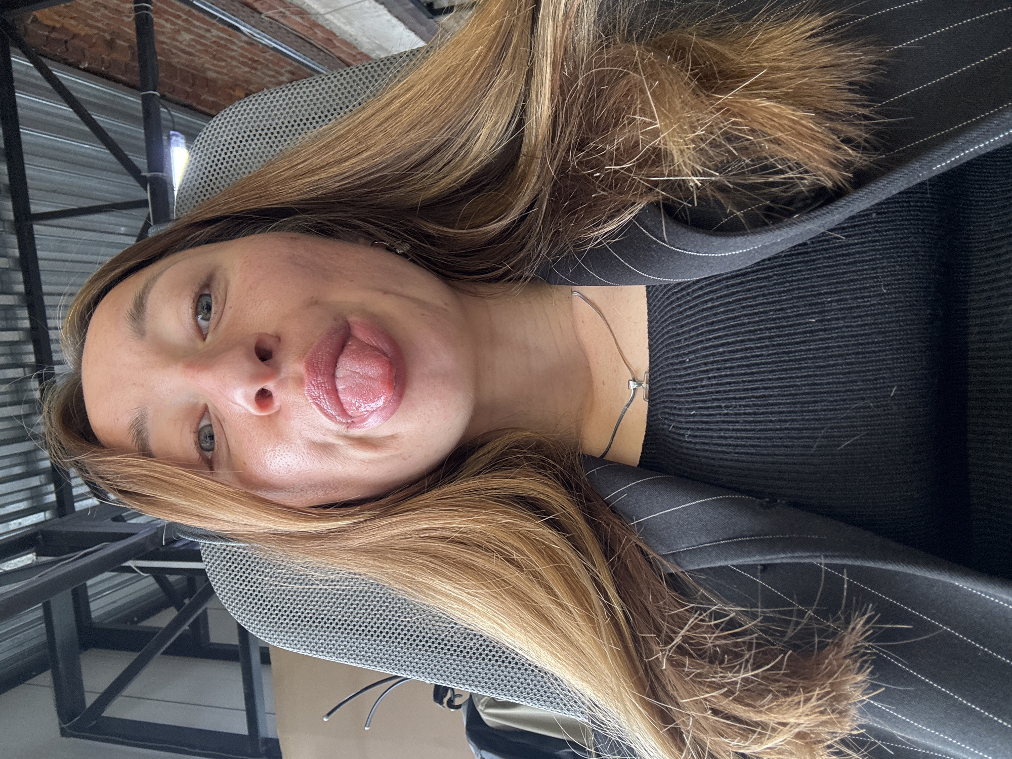 A woman is sitting in a chair. She is wearing a black shirt with a gray pinstripe blazer over it. She has long blond hair and is sticking her tongue out. The environment appears to be an office setting with metal framing. There is no indication of the location being Chaikovsky, Russia.