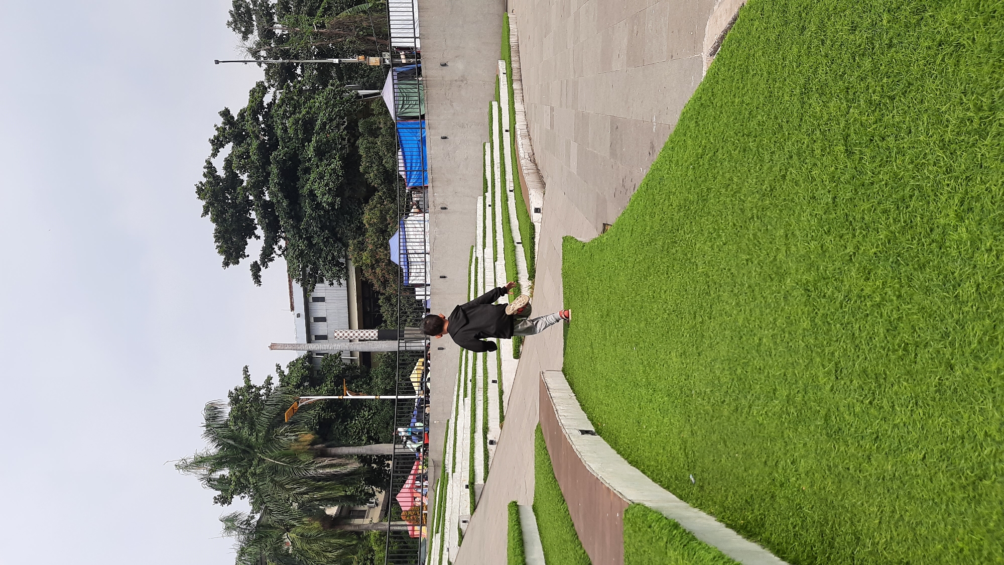 A young child, seen from the back, runs across a stepped grassy area in an outdoor urban public space. The child is wearing a dark hooded sweatshirt, grey pants, and red shoes. The grassy area features a modern tiered design with white concrete borders and wooden accents. Beyond the grass and concrete, a black metal fence encloses a section of the park where several tents of various colors, including blue and green, are set up. In the background, palm trees and dense foliage are visible, along with a multi-story building and a tall, cylindrical concrete structure that appears to be part of a tower or chimney. The sky is overcast. The scene is consistent with an urban park or recreational area in Bandung City, Indonesia, possibly an event space or a public gathering point.