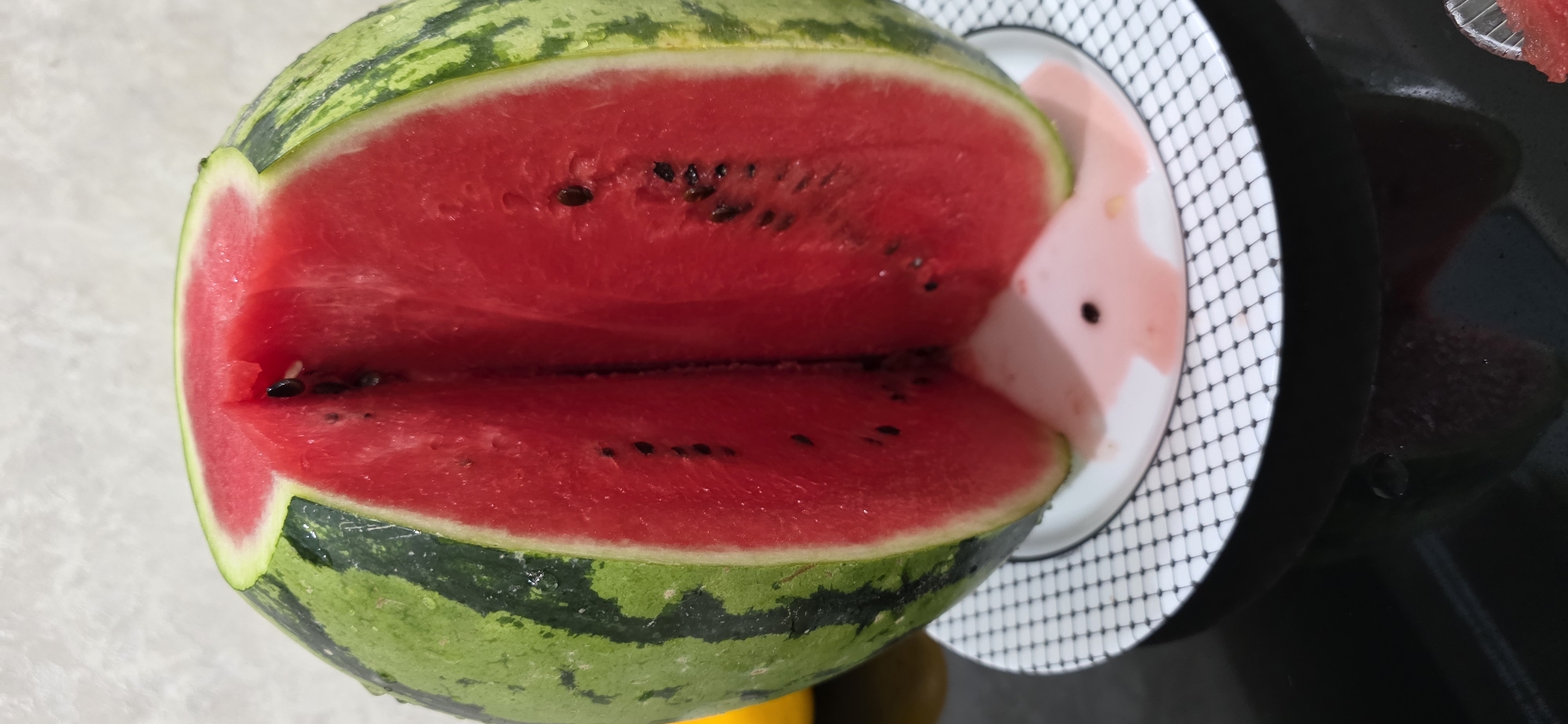 A partially cut watermelon sits on a plate with a black and white checkered rim. The watermelon is round with green rind and a bright red interior, showing its seeds. A portion of the top has been removed, creating a V-shaped opening. Pink juice has pooled on the plate beneath the cut. The background is a textured, light-colored wall. The scene is indoors, likely on a table, with a dark, reflective surface visible below the plate. No people are present, and no specific location in Inozemtsevo, Russia, can be determined from the image.