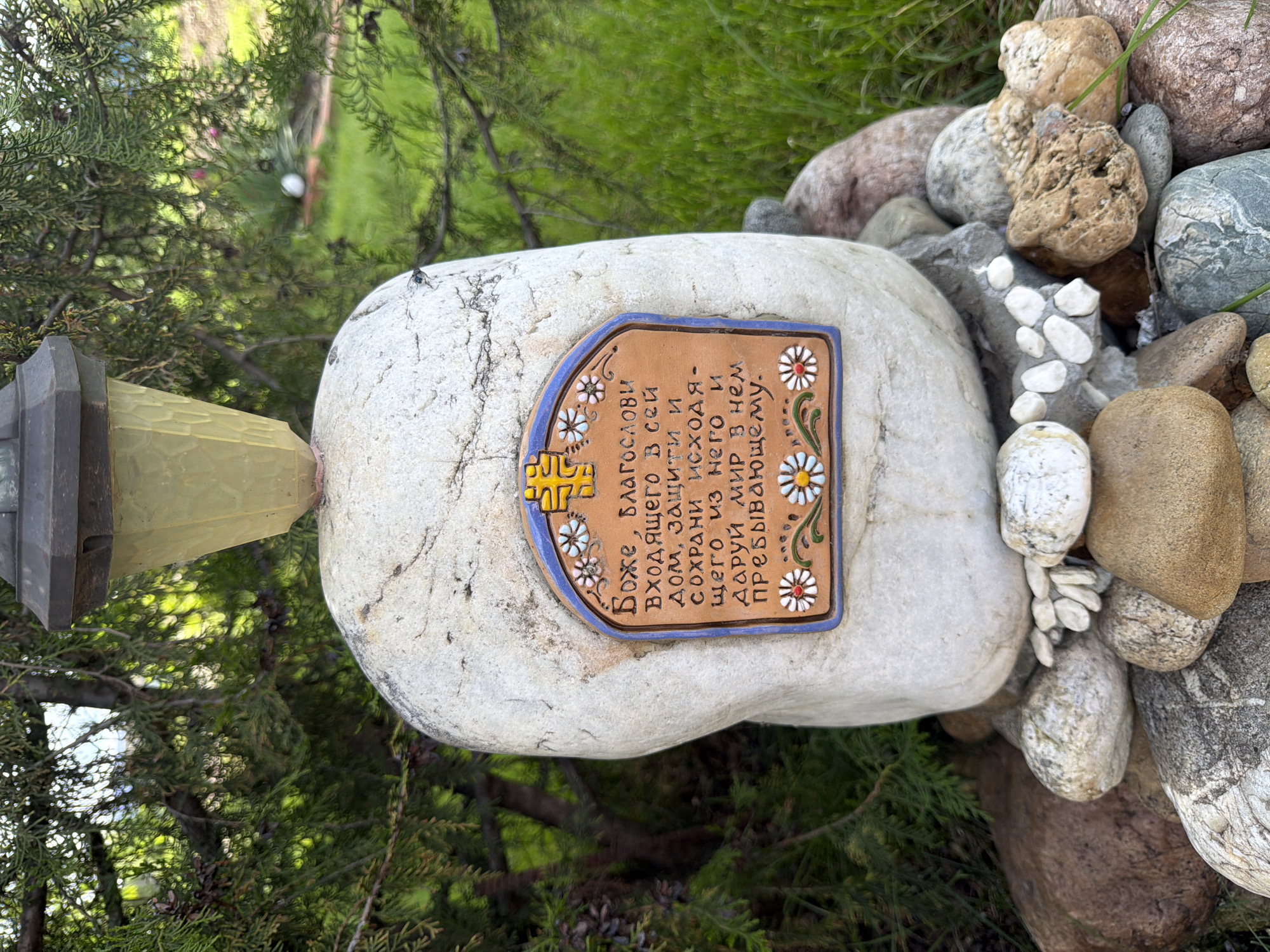 An outdoor scene features a large, smooth, light-colored stone, rotated 90 degrees clockwise in the image, with a decorative ceramic plaque attached to its surface. The stone is surrounded by an assortment of smaller natural rocks of varying sizes, colors, and textures, along with green foliage, possibly from juniper bushes or similar evergreens, and some grass. A small, dark-colored fly is visible on the large stone. Part of a yellow, textured glass lantern or light fixture is visible on the far left.
The ceramic plaque is rectangular with a rounded top, framed by a blue border. It features a cross-like symbol at the top left and decorative white and red daisy-like flowers with green stems along its sides. The plaque contains text in Cyrillic, which reads: "Боже, благоСЛОВИ входящего в сей дом, защити и СОХРАНИ исходящего из него и ДАРУЙ МИР в нем пребывающему." This translates to: "God, bless him who enters this house, protect and preserve him who leaves it, and grant peace to him who abides in it." The scene appears to be set in a garden or a landscaped outdoor area during daylight hours, conveying a sense of tranquility and a spiritual blessing for a home.