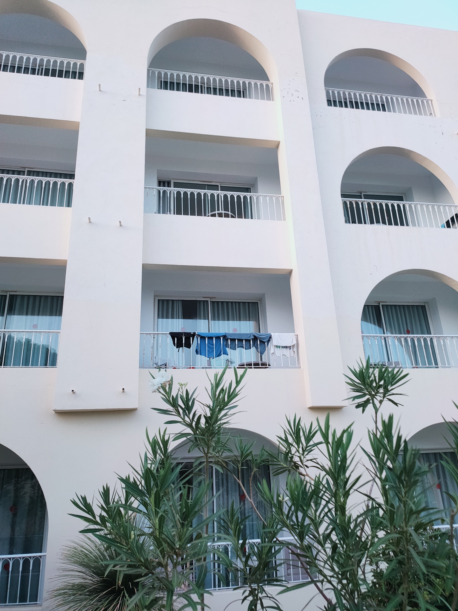This is a medium-angle, outdoor, eye-level shot of a white, multi-story building with arched balconies. The building appears to be a hotel or residential complex. On one of the balconies, clothes are hung to dry, including a black item and a blue towel, as well as a white t-shirt with lettering. Green foliage from trees and bushes is in the foreground, partially obscuring some of the lower balconies. The sky is a pale blue. The architectural style suggests it could be in a Mediterranean region. This building is located in Nabeul, Tunisia.