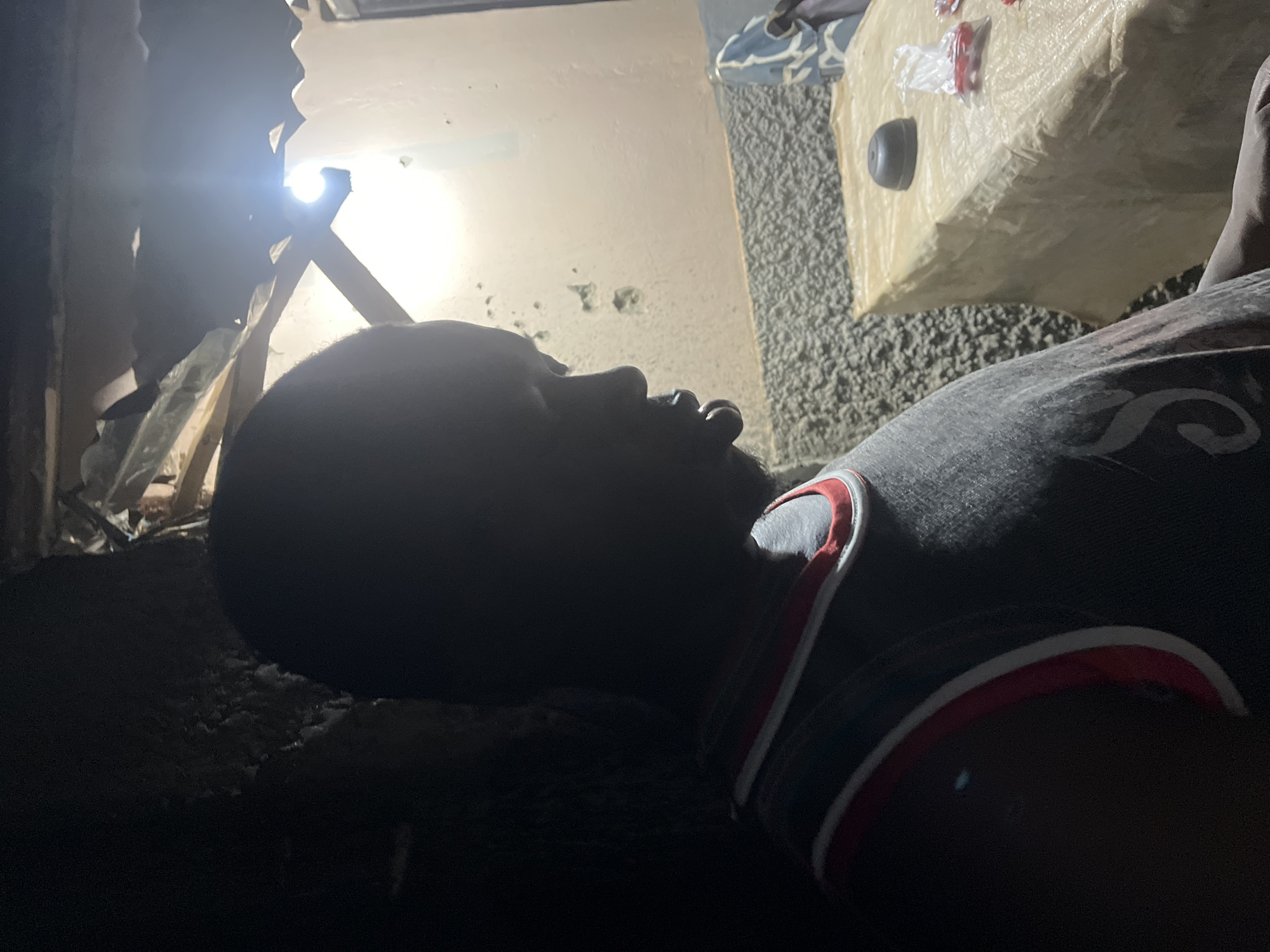 An individual, appearing to be an adult or late teen of African descent, is positioned in profile, facing right, within a dimly lit indoor space in the city of Abuja, Nigeria. The person is heavily silhouetted against a lighter background due to strong backlighting from an exposed, unshaded light bulb suspended from the ceiling. They are wearing a dark gray sleeveless top or jersey with red and white trim around the neckline and armholes, and a partial letter "S" or similar graphic is visible on the chest. The ceiling structure above the light bulb displays exposed wooden beams and corrugated roofing material, along with draped plastic sheeting. To the right of the individual, a table is present, covered with a light-colored, possibly plastic, material. On the table, a dark, round, cap-like object is visible, along with several small, red-wrapped items contained within a clear plastic bag. The background wall is light beige, with a textured finish on the lower section and some dark marks or holes on the upper part. A blue and white patterned fabric object is partially visible in the far right background.