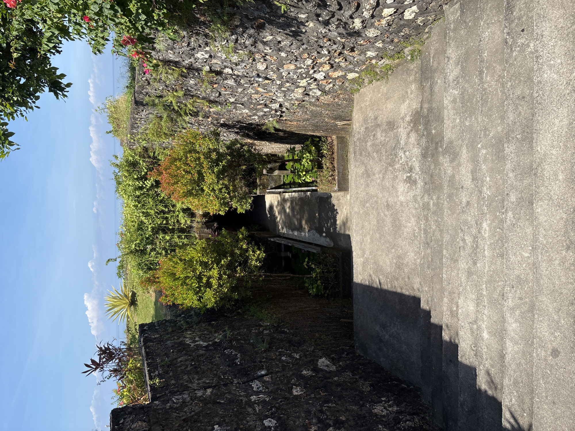 The image depicts a descending concrete pathway flanked by stone walls in Kuta Selatan, Indonesia. The walls are constructed of dark, rough-hewn stones, possibly volcanic rock. Vegetation, including trees and shrubs, grows along and above the walls. At the end of the pathway, a view of the sky and the ocean is visible. A single yellow tree is near the vegetation. The lighting suggests it is daytime with a clear sky. There are no people visible in the image.