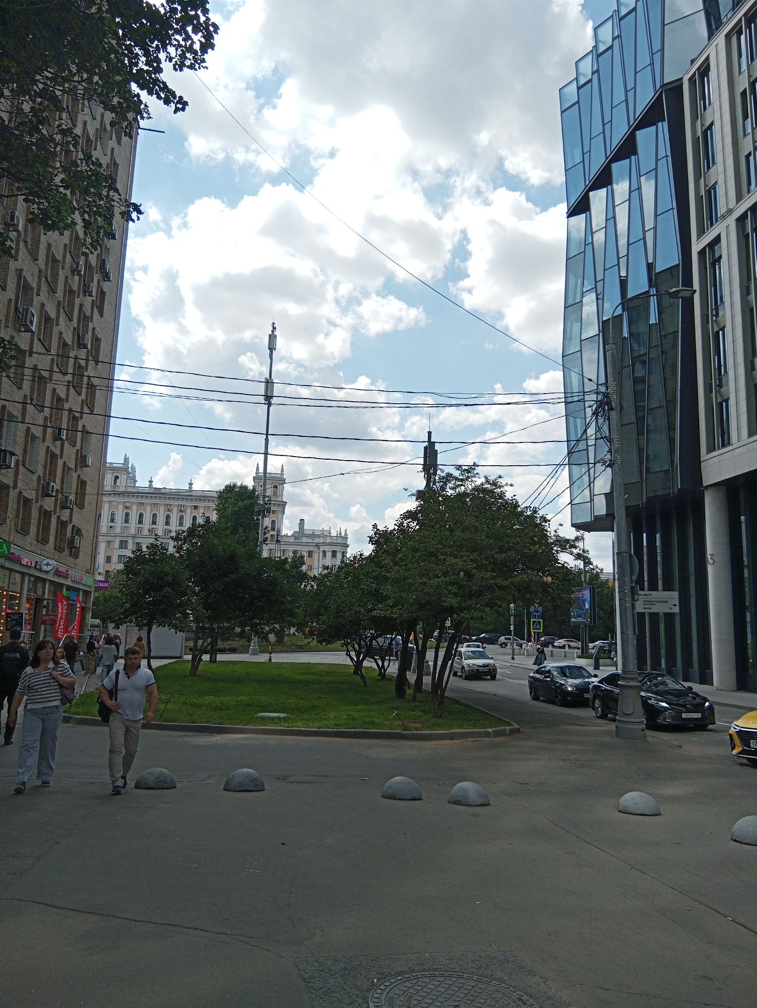 In an urban environment, a street scene unfolds under a cloudy sky. On the left, a tall, beige brick building with numerous windows and air conditioning units lines the sidewalk, where pedestrians are walking. To the right, a modern building with a striking glass facade dominates the view. Between these two structures, a street is visible with several cars, including a black sedan and a white SUV. A small patch of green grass with trees sits in the foreground, bordered by a street. Overhead, a network of power lines crisscrosses the sky. The street is marked with various traffic signs and bollards.