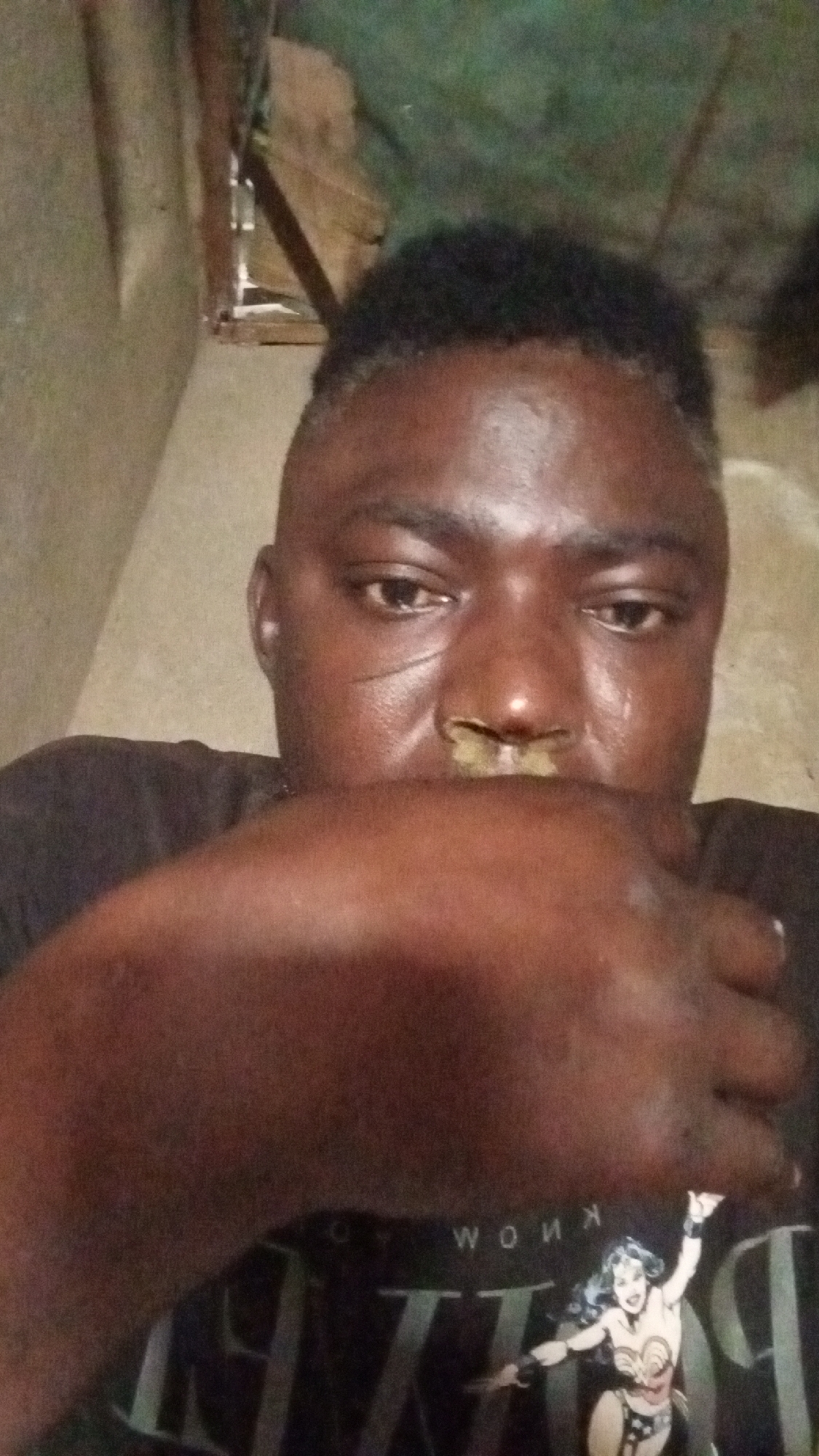 A dark-skinned person, likely male, is captured in a close-up portrait, looking directly into the camera. Their left hand (viewer's right) covers their mouth and lower nose, with fingers slightly curled. The person's eyes appear moist or teary, conveying a sense of sadness, introspection, or distress.
Distinct facial details include a visible scar or mark on their left cheekbone (viewer's perspective) and a yellowish-greenish substance on their nose, just below the nostrils. The person has short, dark, textured hair. They are wearing a dark t-shirt featuring a graphic of Wonder Woman, with the character prominently displayed. Above the character, the word "KNOW" is visible in reversed text, and below, larger letters are present, likely forming part of "WONDER WOMAN."
The background is dimly lit and indistinct, suggesting an indoor environment. Visible elements include what appear to be wooden structures or panels, indicating a simple or possibly unfinished setting. The low light contributes to a somber atmosphere. No specific location or time of day is discernible.