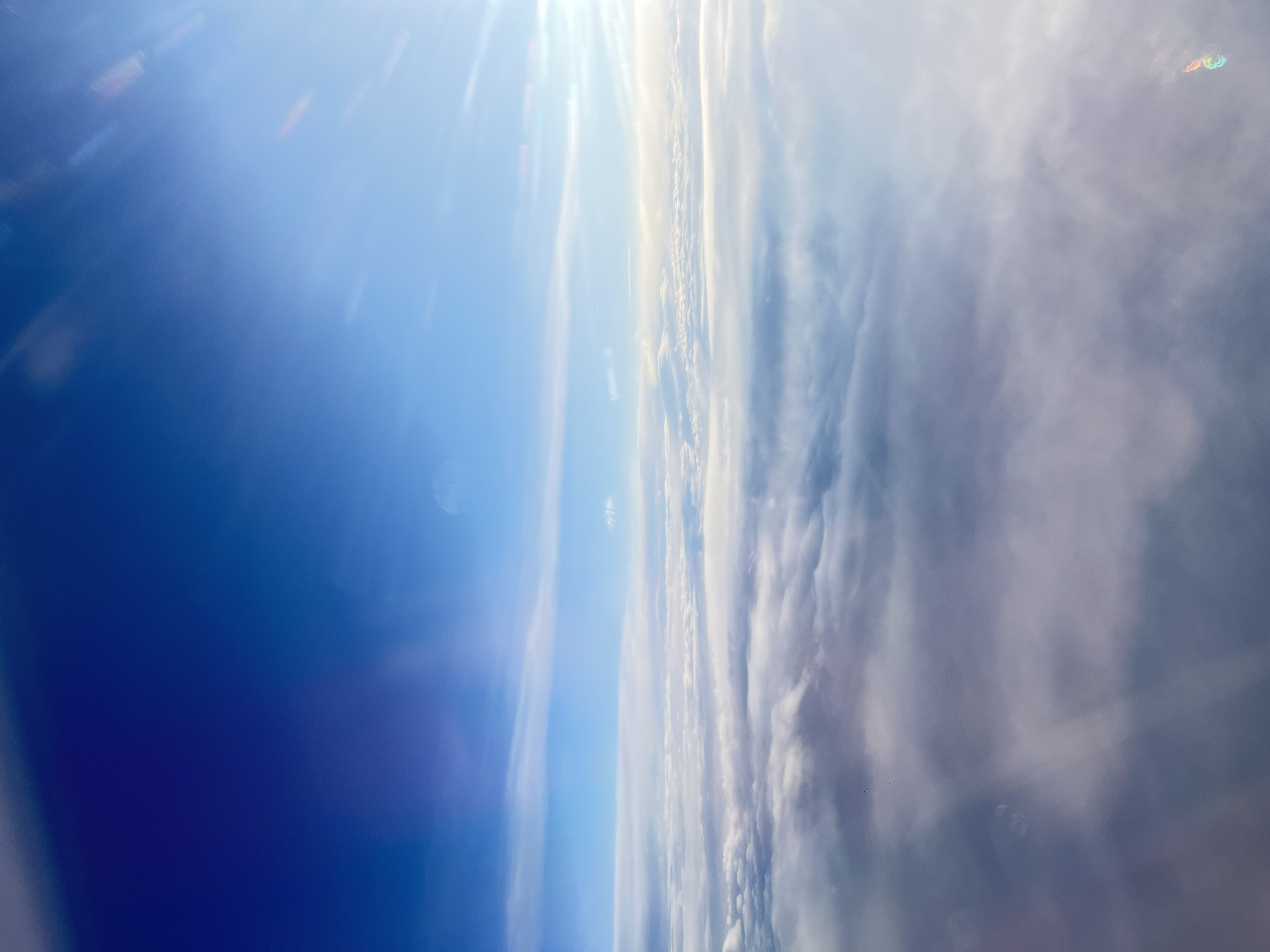 The vertically oriented image captures an expansive aerial view of the sky and clouds, likely taken from high altitude. On the left side, the sky transitions from a deep, rich blue at the bottom to a brighter, lighter blue towards the top. A brilliant white light, identified as the sun, emanates from the top-left corner, causing significant lens flare that streaks across the blue expanse.
The right side of the frame is dominated by a vast, thick bank of clouds, which appear to stretch horizontally across the image. These clouds exhibit a range of tones, from bright white where they are intensely lit by the sun to softer greys in shadowed areas, creating a textured and undulating mass. The boundary between the clear blue sky and the extensive cloud cover is a distinct feature running vertically down the middle of the image.
Several small, subtle colorful specks, consistent with lens artifacts or reflections, are faintly visible, particularly against the clouds and in the upper sky areas. The scene suggests daytime conditions due to the bright sunlight. The overall impression is one of immense scale, tranquility, and isolation, typical of a view above a significant cloud layer from an aircraft. No visible text is present in the image.