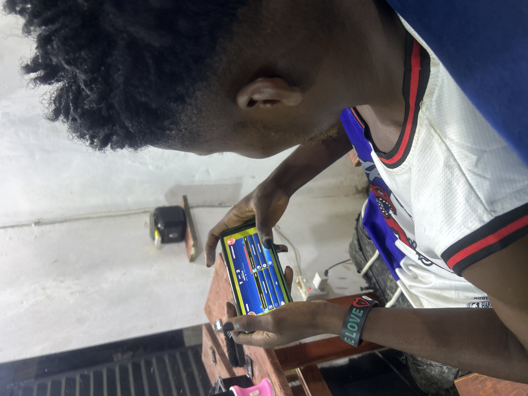 The image captures a close-up, low-angle view of a young person engrossed in their smartphone. The subject, with dark skin and short, dark curly hair, is seen from a side profile, looking down at the phone held in both hands. Their body is angled to the right, wearing a white sleeveless top with black and red trim around the neckline and shoulder, layered with a blue garment. A black wristband with the word "LOVE" in white text and a red heart symbol is visible on their left wrist.
The smartphone screen, brightly illuminated, displays an application with a blue background featuring vertical bars, possibly indicating an audio equalizer, a music production app, or a game interface. Visible text on the screen includes "VICTORY" and the numbers "1 - 2". The person appears highly focused and engaged with the device.
The setting is an indoor environment, with a light-colored wall or surface forming the background. A rustic wooden structure, possibly a table or stand, occupies the lower part of the frame, beneath the person's hands. In the background, on a lighter surface, several indistinct objects can be seen, including a black rectangular item and a small white object with a red light. The lighting is consistent with indoor artificial illumination. The scene depicts a casual moment of interaction with technology, likely in a home or personal space in Ikorodu, Nigeria.