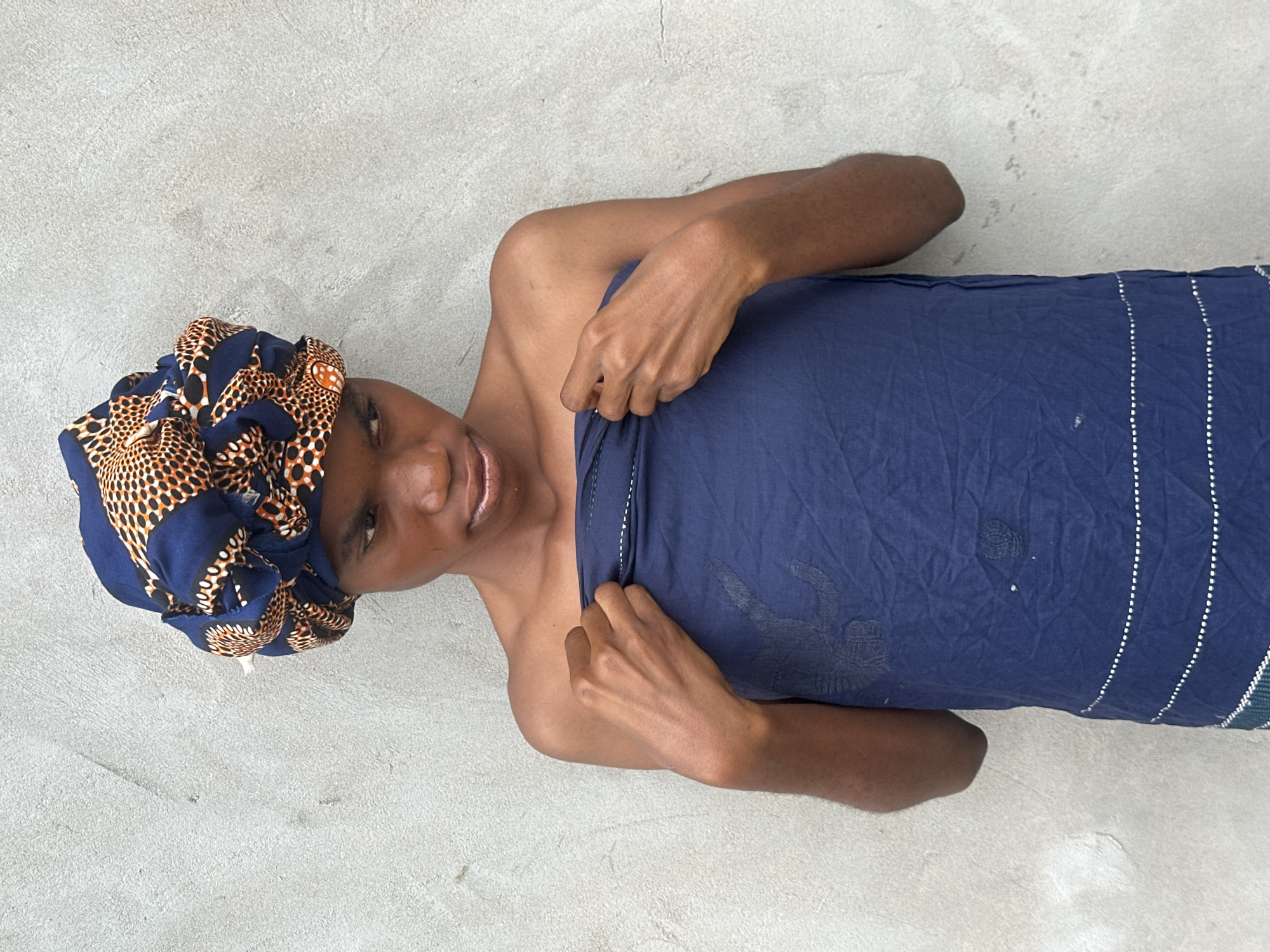 A young person, with dark skin and wearing a vibrantly patterned headwrap, lies on a light grey concrete surface. The headwrap features a design of blue, orange, and black spots and shapes. The person is also draped in a dark blue fabric, likely a wrap or skirt, that covers their lower body and chest. Their left arm is bent at the elbow, with their hand resting on the blue fabric, pulling it slightly. The person's face is turned towards the camera, with an open-mouthed expression that suggests they might be speaking, laughing, or reacting to something. Their eyes are looking slightly to the left. The lighting suggests it is daytime, possibly outdoors, given the light grey concrete setting which is common in many outdoor areas in Badagry, Nigeria. The scene is simple and focused on the individual, with no other objects or people visible. The overall mood is relaxed and informal.
