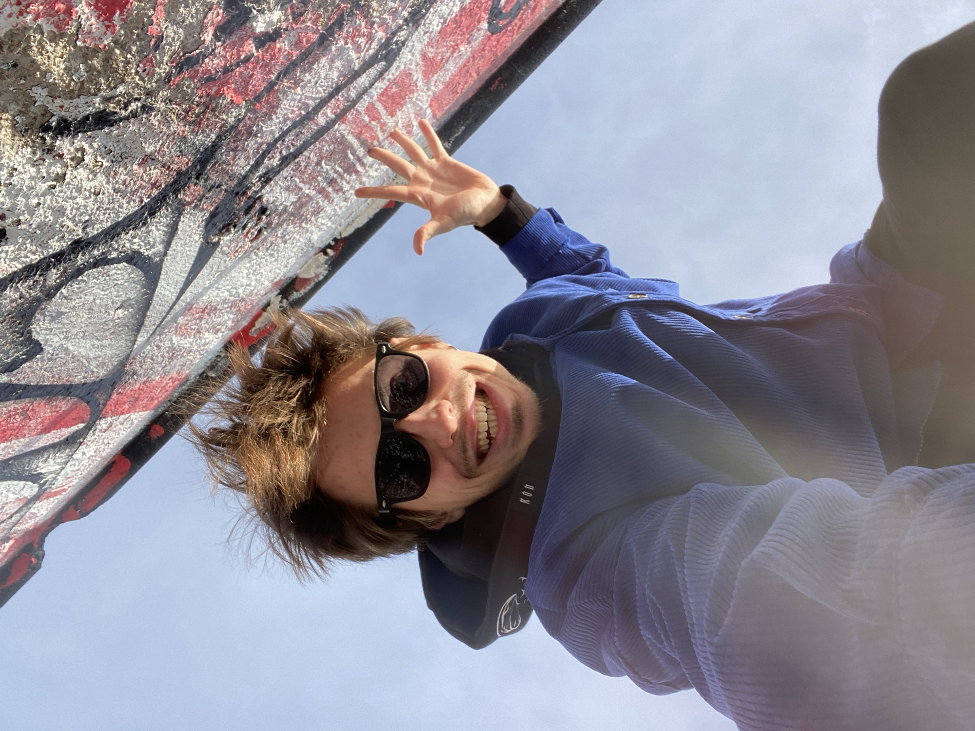 The image is an upward-facing selfie taken outdoors in daylight. A person is centered in the frame, wearing dark sunglasses and a blue corduroy jacket. They have brown hair and are smiling with teeth visible. The person is standing next to a wall covered in graffiti. The sky is visible in the background. There is a street artist nearby in Bar, Montenegro.