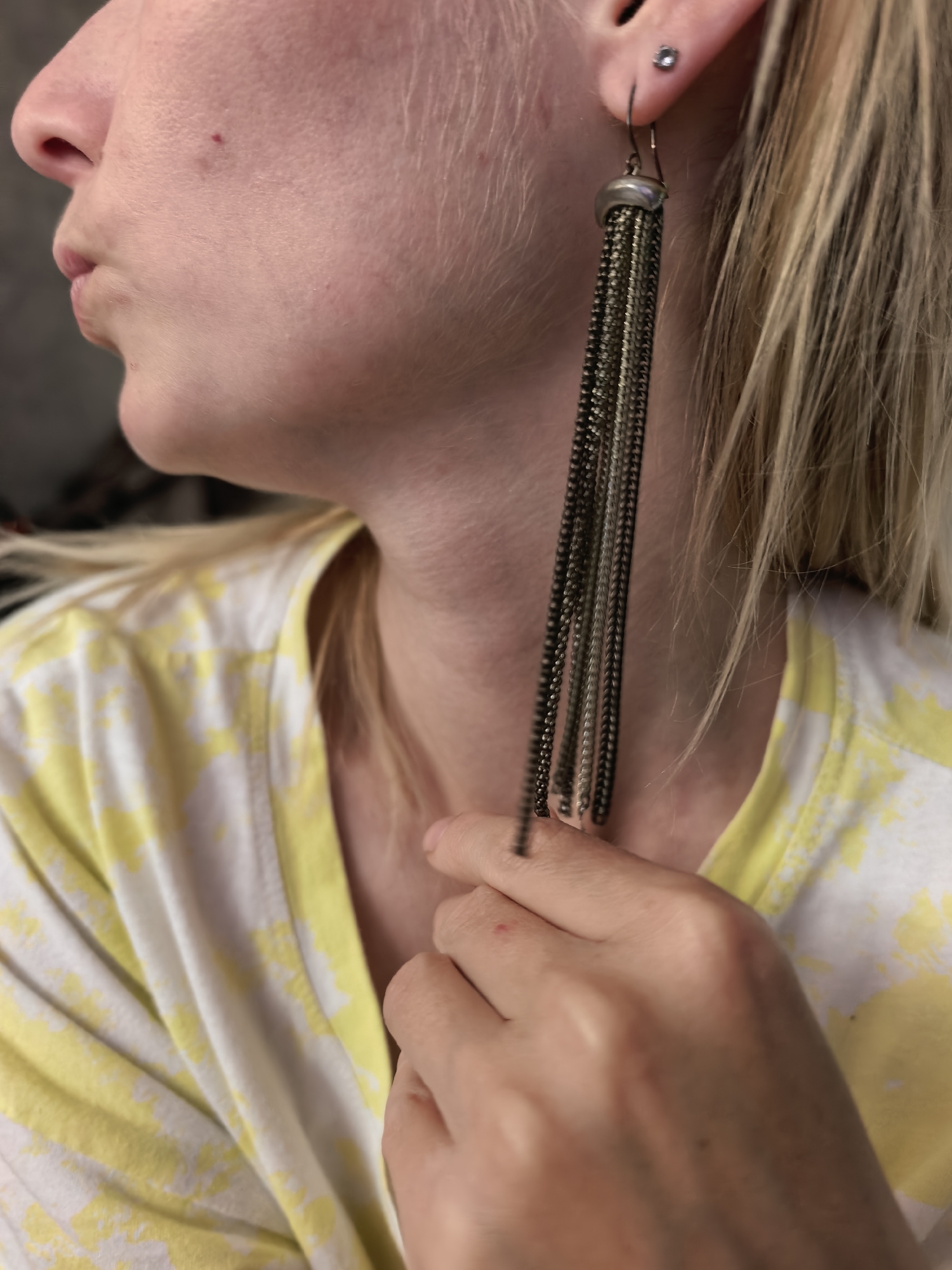 The image shows a close-up of a person's face, ear, and shoulder, with their hand touching their ear and earring. The person has fair skin and blonde hair, and is wearing a yellow and white tie-dye shirt. The earring is a long, dangling chain earring with a metallic top. A small stud earring is visible in the lobe of the ear. There is no indication of the location in the city of Sterlitamak, Russia, as this is a cropped image of a person and their attire.