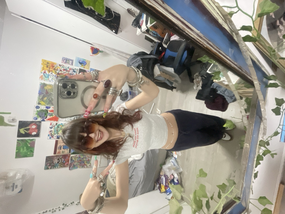 This image captures a mirror selfie of a young female within a room located in Raanana, Israel. The subject, possessing long reddish-brown hair with bangs, wears large dark-tinted sunglasses, a white cropped t-shirt featuring inverted red text, dark blue sweatpants, and white shoes with green socks. She displays numerous rings on her fingers, multiple bracelets on her wrists, and a thin beaded chain around her waist. Her fingernails are long and multicolored. She holds a light-colored smartphone in her right hand to take the photograph, while her left arm is extended, forming a "V" shape with her fingers. A wide smile is evident on her face, with her tongue slightly protruding. The background, reflected in the mirror, reveals white walls largely covered with an assortment of colorful artworks and posters. To the left, a bed with grey bedding is partially visible, with items scattered on the adjacent light-colored floor. To the right, a white wardrobe and a dark office chair draped with clothing are discernible, along with other miscellaneous objects and debris on the floor. Artificial green vines are strategically placed around the mirror frame and on the wall.