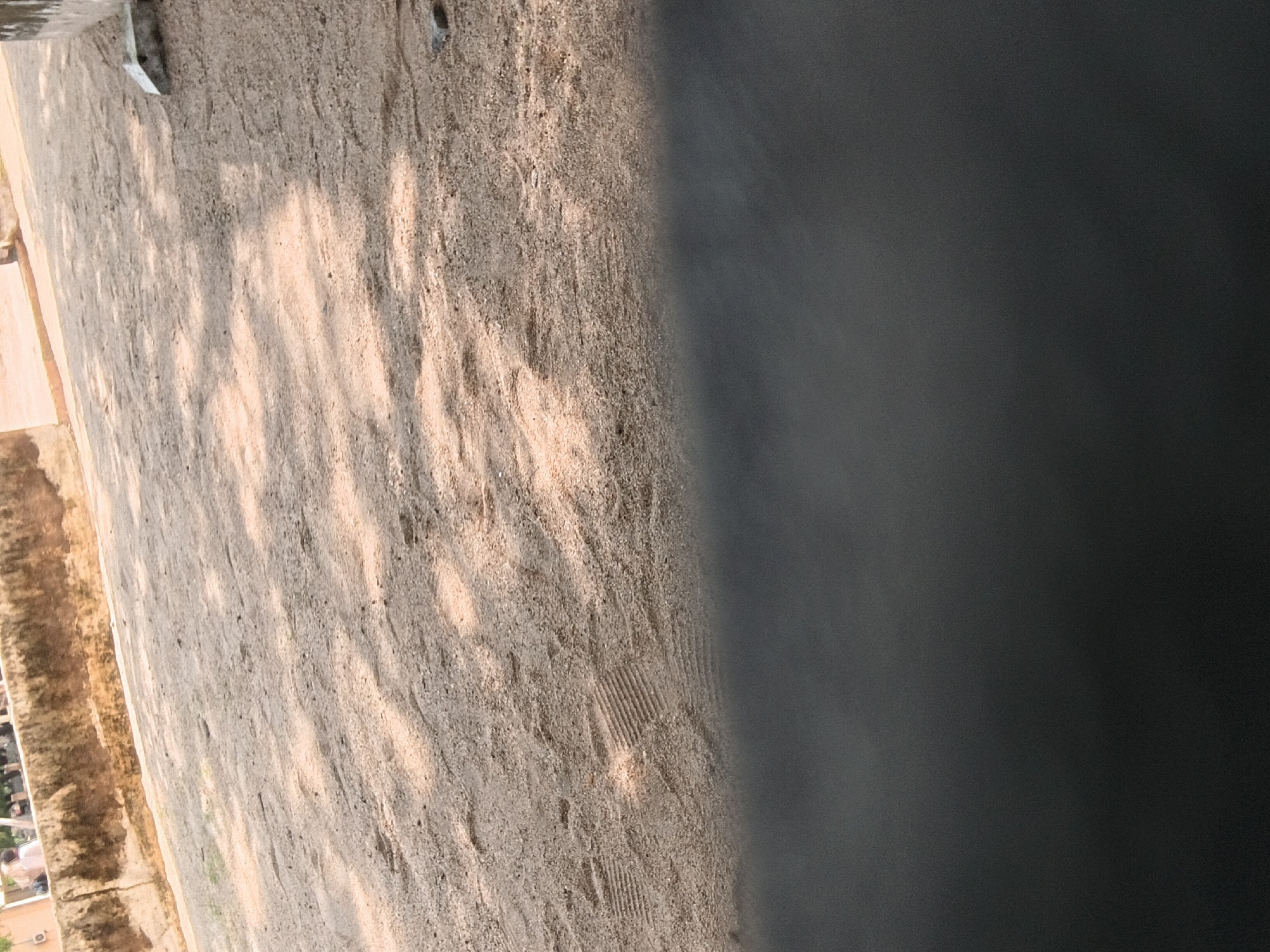 A scene of a sandy area with some footprints and shadows from trees overhead. In the background, a low wall runs across the image, with a few figures visible behind it. A small concrete block sits on the sand near the wall on the right side of the frame. The lower portion of the image is obscured by a dark, blurry foreground. The location is identified as Jalingo, Nigeria.