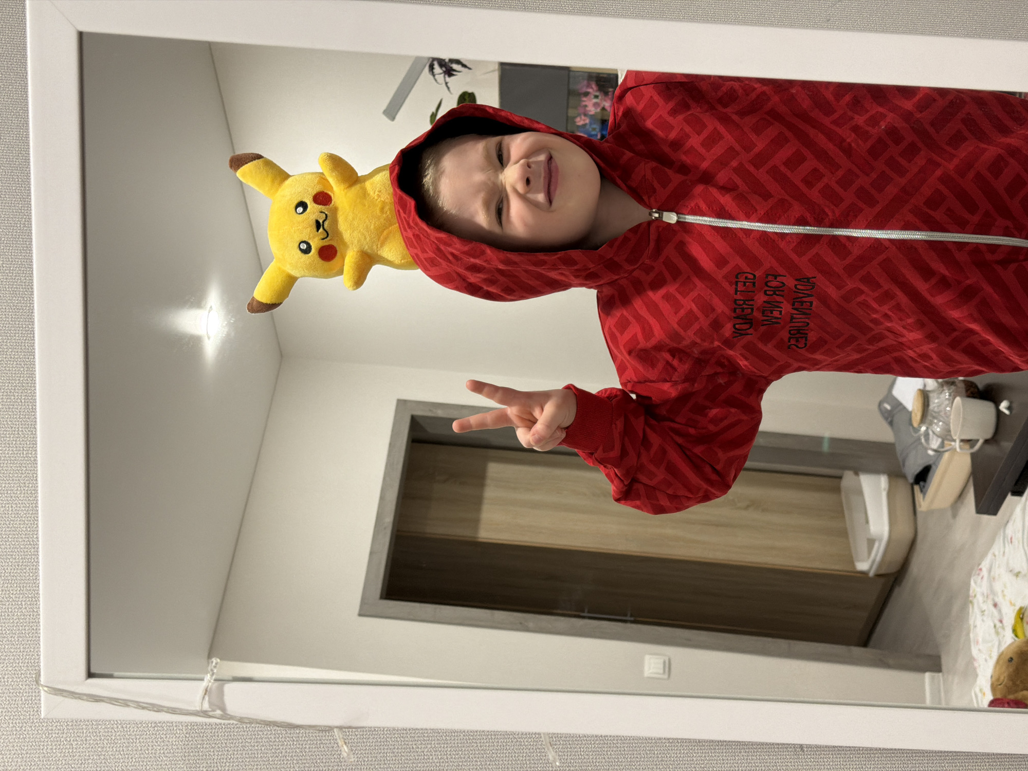 A child in a red, patterned zip-up hoodie and a Pikachu plush are reflected in a mirror with a white frame. The child is making a peace sign with their hand. The mirror is hanging in a room with neutral-toned walls. A doorway with a light brown door and grey frame is visible in the reflection. A small portion of a bed and bedside table with various items on it is visible as well. The location is Засечное, Russia.