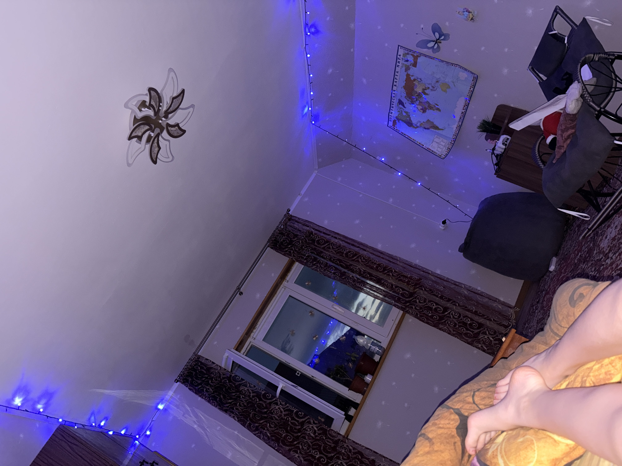 The image depicts an interior scene, likely a bedroom, illuminated by blue string lights. A window with patterned curtains is visible, showing a nighttime view with faint lights. A world map is mounted on the wall, along with decorative elements like a butterfly. Various pieces of furniture, including a beanbag chair and rocking chairs, are present. The overall ambiance is cozy and dimly lit, with the blue lights creating a distinct atmosphere. The image appears to have been taken in Leninogorsk, Russia, though specific details about the city are not discernible from the visual content alone.