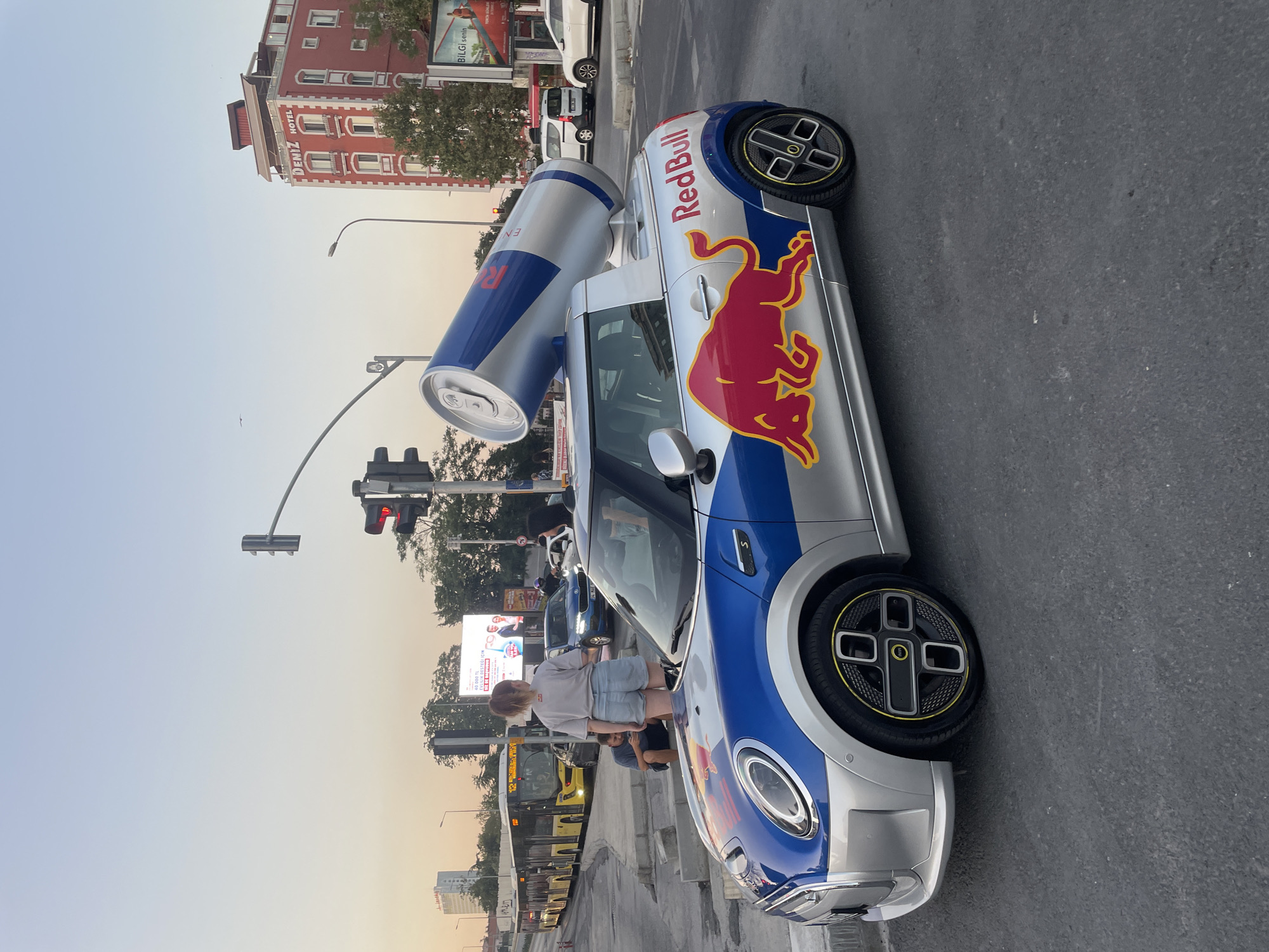 The image shows a customized Mini Cooper pickup truck, branded with Red Bull logos and featuring a large, oversized Red Bull can mounted on its bed, positioned on a city street in Kadıköy, Turkey. The vehicle is silver and blue, with a prominent Red Bull bull graphic on its side. The truck is stopped at a traffic light intersection. In the background, a yellow bus is visible, as are several buildings, including a red building with "DENİZ HOTEL" signage. A few people are present near the Mini Cooper, including a young woman standing and looking towards the bus, and a man sitting on a concrete step. The sky is clear and light blue, suggesting it's either early morning or late afternoon. Traffic lights are illuminated, with one showing red. The overall scene depicts a promotional event or display in an urban environment.