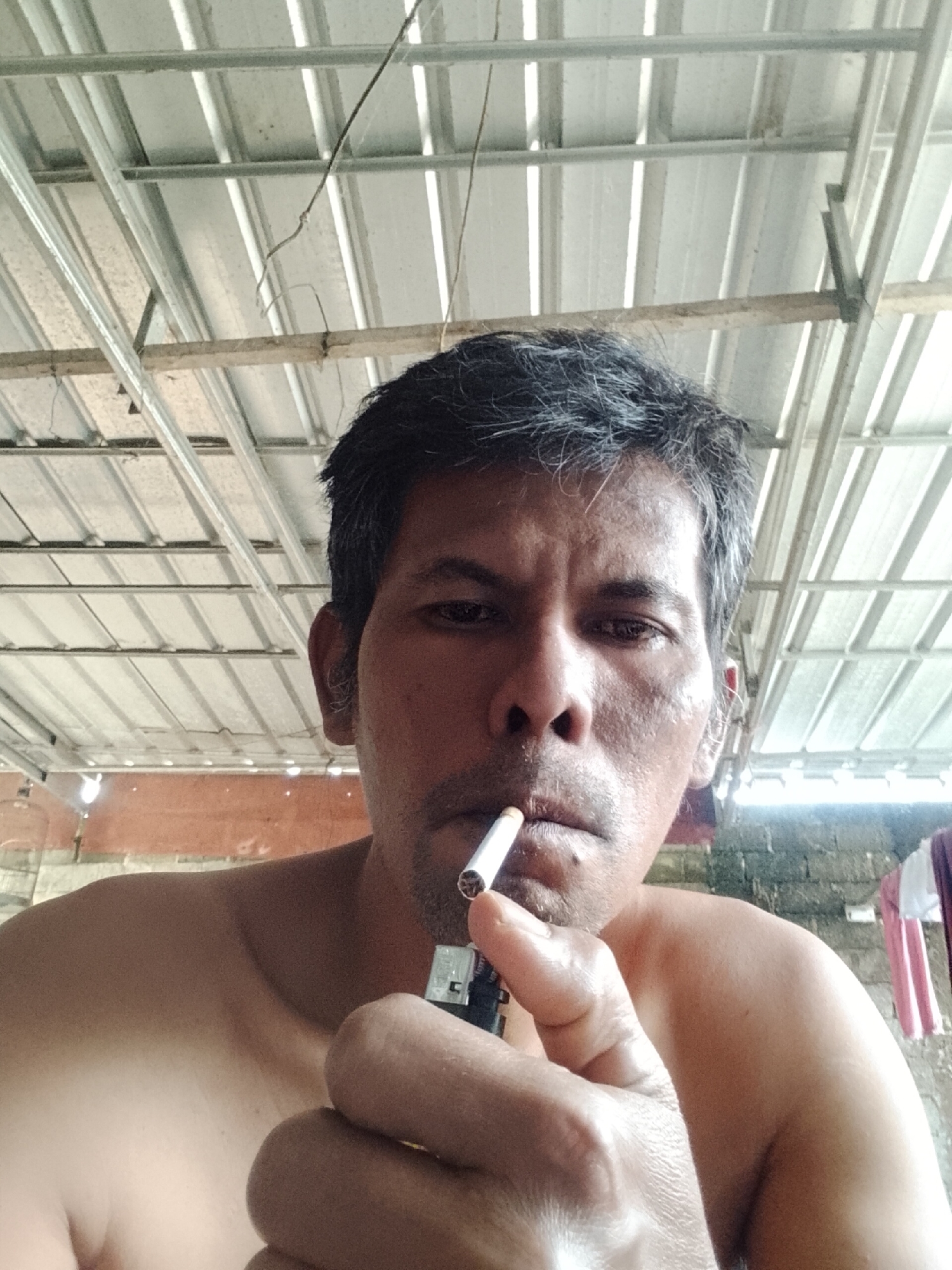 A man is holding a lit cigarette and a lighter to his face, in an outdoor setting with a corrugated metal roof overhead. The man has dark, short hair and appears to be of Asian descent. He is wearing a light-colored shirt that is partially visible. In the background, there is a brick wall and some laundry hanging on a line. The overall scene suggests a casual, informal environment. The location is Padang Dalom, Indonesia.