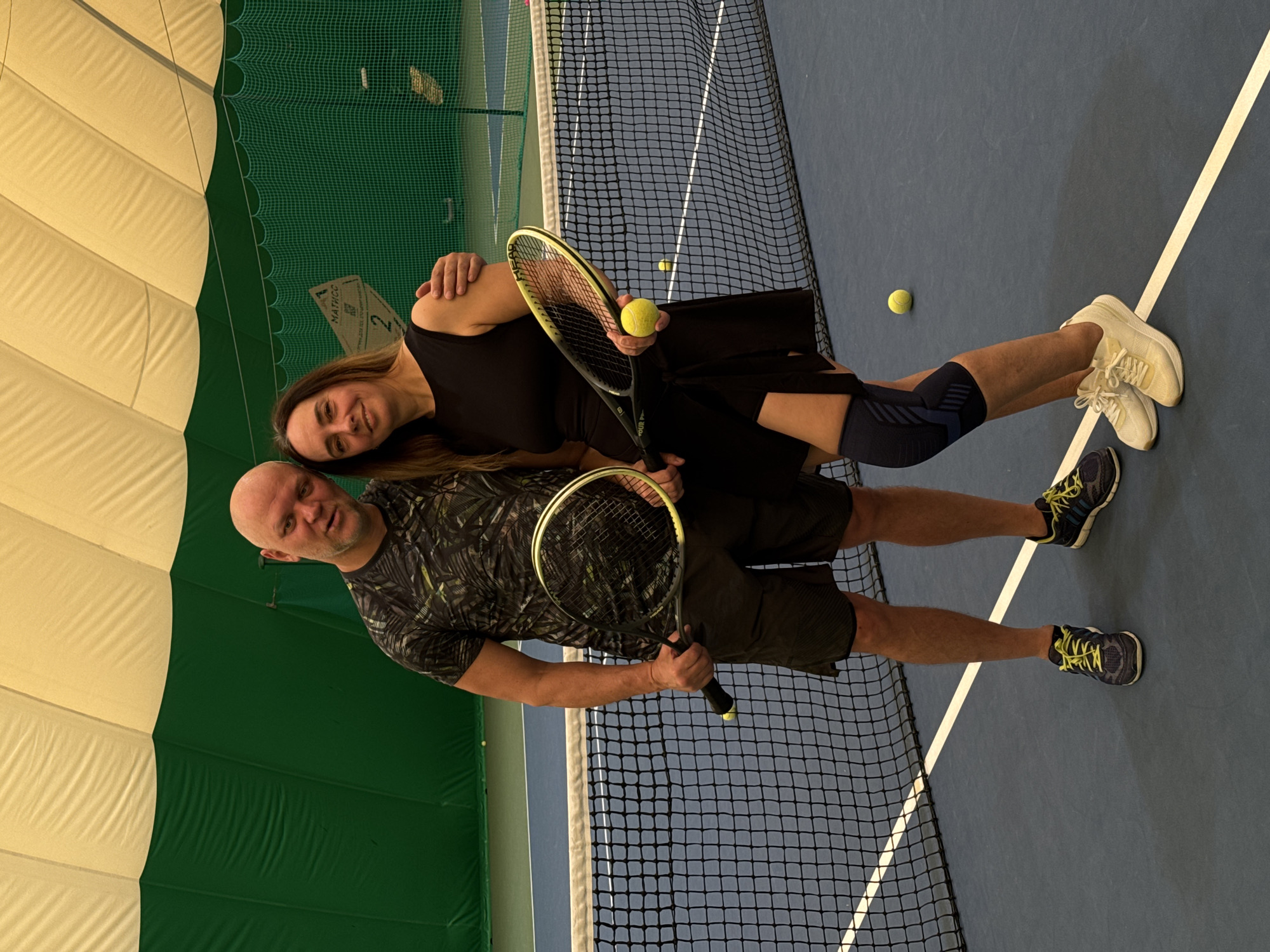 In a rotated, eye-level, indoor shot, a man and a woman are standing on a blue tennis court with a net in front of them, likely in Nizhny Novgorod, Russia. The man has a shaved head and is wearing a patterned t-shirt and shorts, holding a tennis racket in his right hand. He looks towards the camera. The woman, who has long brown hair, is wearing a black dress and knee pad. She rests her left arm on the man's shoulder. She is holding a tennis racket in her right hand, with a tennis ball in the strings. In the background, there are tennis balls on the court. The walls are partially visible, with a green and white structure visible on the left. The tennis court lines are white.
