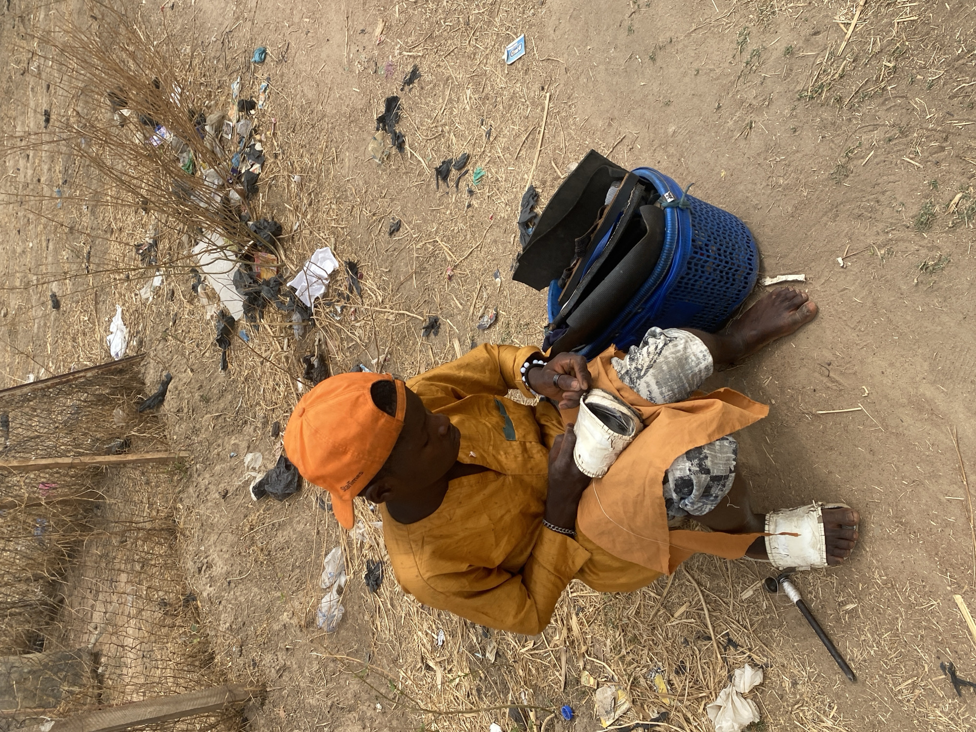 The image shows a man sitting on the ground in Rido, Nigeria. He is wearing an orange cap and shirt, and patterned pants. He appears to be mending shoes or sandals. A blue basket sits behind him, containing multiple pairs of shoes. The ground is dirt, strewn with trash and dried brush. Shoe repair tools lie next to his feet. One of his feet is bandaged.