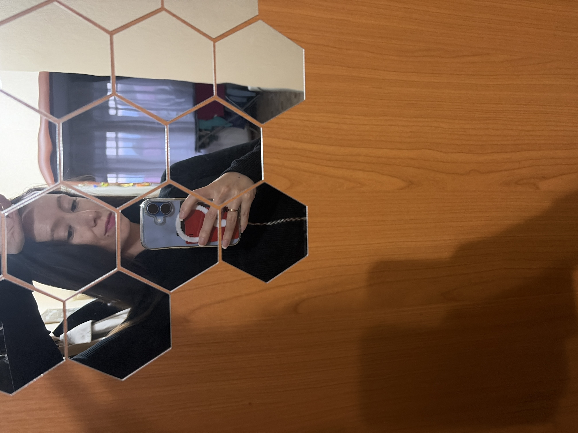 The image shows a woman taking a selfie in a mirror. She is wearing a black top and has long dark hair. She is holding a smartphone in her right hand, and her left hand is resting on her head. The mirror is decorated with hexagonal tiles, and the background shows a room with furniture and windows. The image does not provide any information about the location in the city of Япрыково, Russia.
