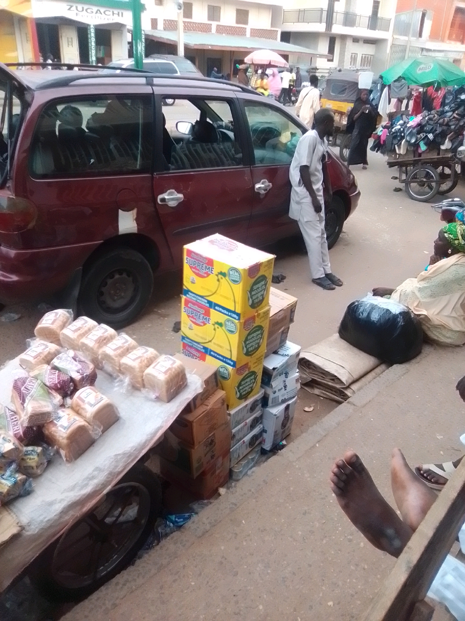 The image captures a street scene in Arawa, Nigeria, with a focus on a market area. In the foreground, several loaves of bread are neatly arranged on a table, wrapped in plastic. To the right of the bread, stacked yellow boxes labeled "SUPREME NOODLES SUPREME TASTE" are visible. A dark red minivan with its rear hatch open is parked prominently in the center of the frame. Several individuals are present in the scene. A man in a light-colored traditional outfit stands near the open minivan door. Further back, a woman in a dark headscarf walks with a load on her head, and another person, possibly a woman, is seated on the ground next to a large black plastic bag. A cart filled with clothing and accessories is positioned to the right, with a green umbrella providing shade. The environment appears to be a bustling street with buildings lining the background. The ground is paved, and debris is scattered in places.