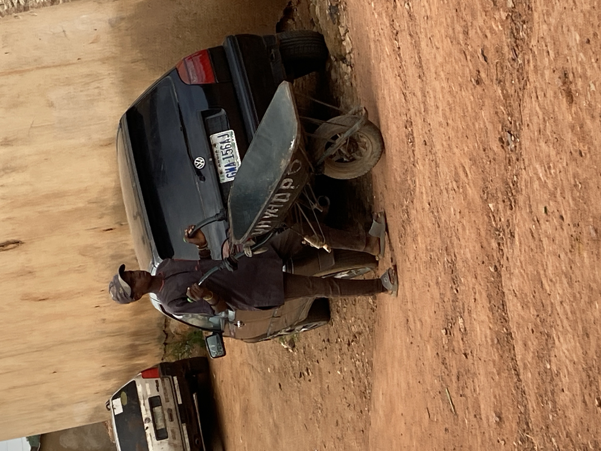 In Bukuru, Nigeria, a young boy stands beside a black Volkswagen Golf hatchback, holding the handles of a wheelbarrow. He is wearing a dark long-sleeved shirt, dark pants, a baseball cap, and light-colored sandals. The wheelbarrow is positioned in front of the car. To the left of the Volkswagen, a white SUV is visible. The ground is covered in reddish-brown dirt with scattered pebbles. The background features a light brown, textured wall. The scene appears to be on a street or an open area.