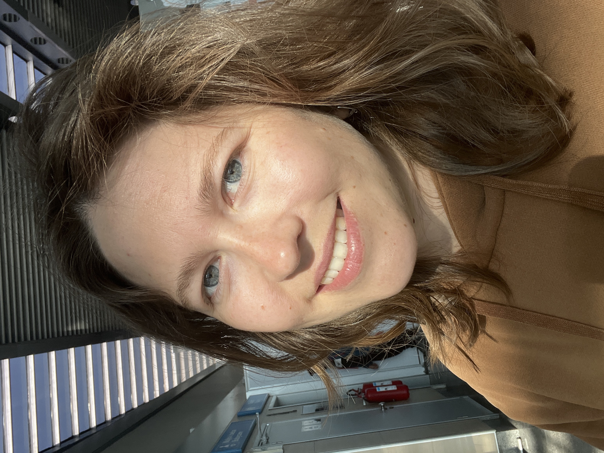 A medium-shot, slightly rotated photograph features a young woman with light skin, blue eyes, and light brown hair. She is smiling, revealing straight, white teeth. She wears a brown top. The background shows a metallic, slatted structure suggestive of an indoor or covered space. Lower in the frame, there's a glimpse of a gray surface, a small red object, and blue rectangular items. The photo was taken indoors. There is no conclusive evidence in the image to establish that this photograph was taken in Podgorica, Montenegro.