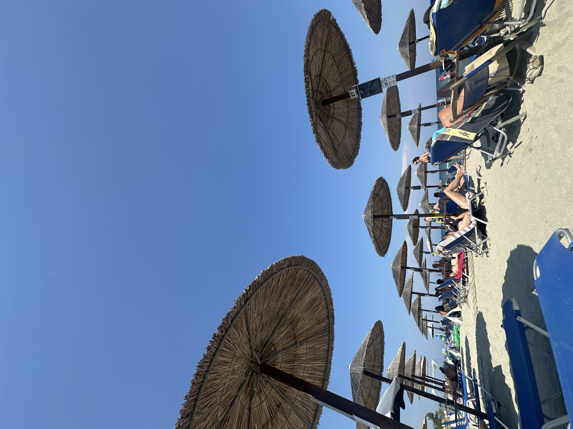 An outdoor, eye-level, wide shot shows a beach scene in Greece under a clear blue sky. In the foreground, several blue lounge chairs are unoccupied, with one in the lower left and another in the lower right corners of the frame. In the mid-ground, rows of beach umbrellas with thatched tops provide shade for numerous people lounging on beach chairs. Some individuals are sunbathing, while others are sitting or talking. The sand is light-colored and appears well-trodden. The background shows the calm blue sea. The overall impression is one of a relaxing summer day at the beach.