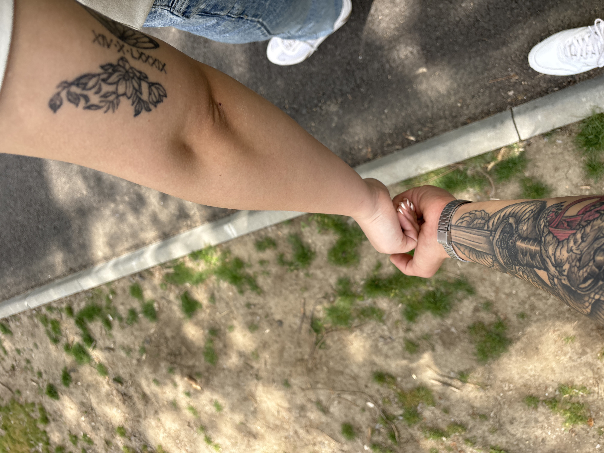 The image, taken from an overhead perspective, depicts two individuals holding hands outdoors, likely in Plovdiv, Bulgaria. The bright daylight is evident from the shadows cast on the ground. The scene shows a paved or asphalt surface on the left, bordering a concrete curb. Beyond the curb, the ground transitions to a dry, light brown dirt area with patches of green grass.
The arm on the left, lightly tanned and bare, extends from the upper left corner. It features a black-ink tattoo on the inner forearm, showcasing a design of blooming flowers and leaves. Above this, a line of Roman numerals reads "XIV-X-LXXXXX". A glimpse of white clothing and blue denim jeans, along with a white sneaker, can be seen in the upper left, indicating the attire of this person.
The arm on the right is heavily adorned with a detailed black and grey full-sleeve tattoo, depicting intricate patterns and what appears to be a sword or a mythical creature. A silver-toned metal watch is worn on the wrist. The hand holding the other has neatly manicured nails, possibly with white tips. The intertwining of their hands suggests a close connection or shared moment.