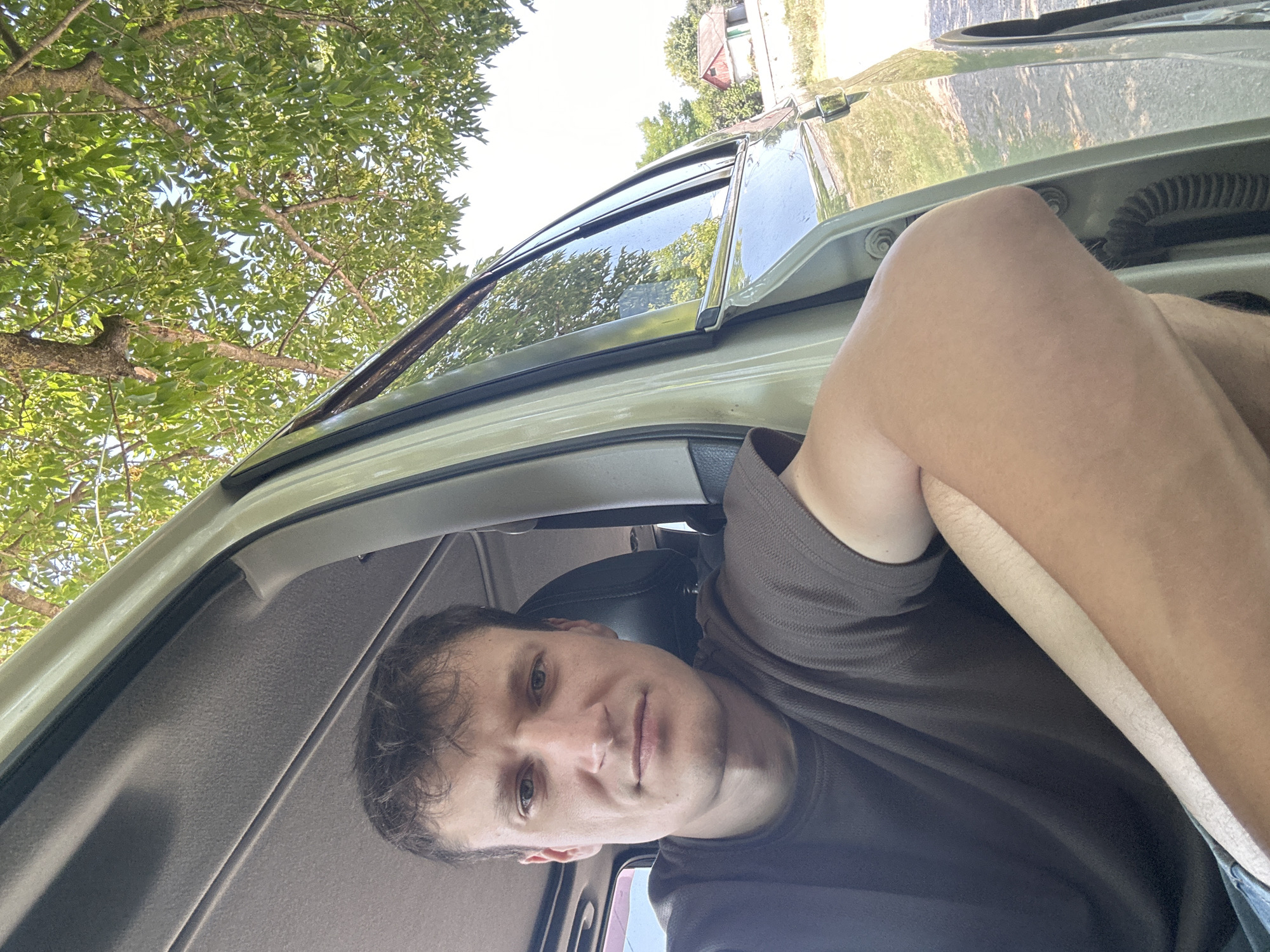 A man with short, dark, messy hair and a serious expression sits in the driver's seat of a light-colored vehicle. He is wearing a dark brown t-shirt and his left arm is resting on the open door, with his elbow bent. The background outside the car shows a bright sky with green leaves from a tree overhead, and in the distance, a small building with a red roof is visible. The image is a close-up, selfie-style shot taken during the day. The location is specified as Makiv, Ukraine.