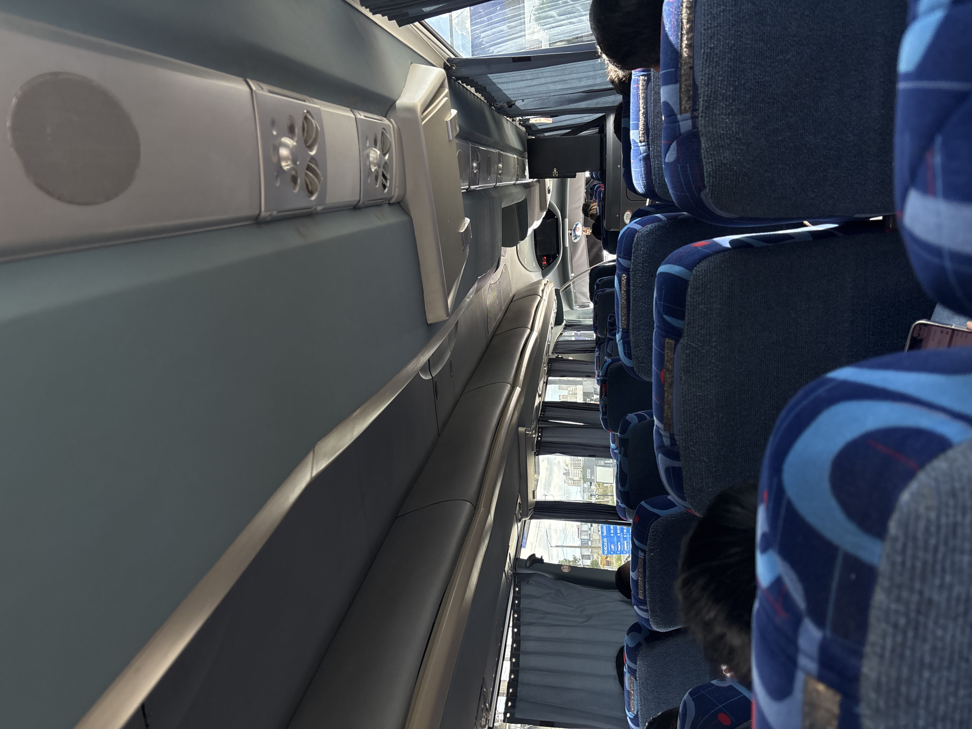 The image presents the interior of a passenger bus in San Bernardino Tlaxcalancingo, Mexico, observed from a rearward perspective looking towards the front. The bus is equipped with multiple rows of blue seats featuring grey fabric on the main backrest and seat cushion, complemented by blue patterned fabric with light blue swirling designs on the headrests and side bolsters. Several passengers occupy the seats, visible primarily from the backs of their heads.
Overhead, a light grey ceiling incorporates recessed lighting and ventilation units. A central panel along the ceiling houses individual air vents, reading lights, and a circular speaker grille. Luggage racks are integrated above the windows on both sides. Light blue curtains are partially drawn over the windows.
Through the right-side windows, an exterior scene is visible. A prominent blue and white sign, consistent with a toll booth or fare collection point, displays text in Spanish: "VEHICULOS - TARIFA" (Vehicles - Fare), followed by a list of monetary values, including "$ 20.00", "$ 30.00", "$ 60.00", and "$ 130.00." Beyond the sign, multiple multi-story buildings are discernible against a bright sky, indicating an urban or semi-urban setting. No explicit actions or interactions between individuals are visible, though a passenger in the immediate foreground on the left side appears to be holding a mobile phone.