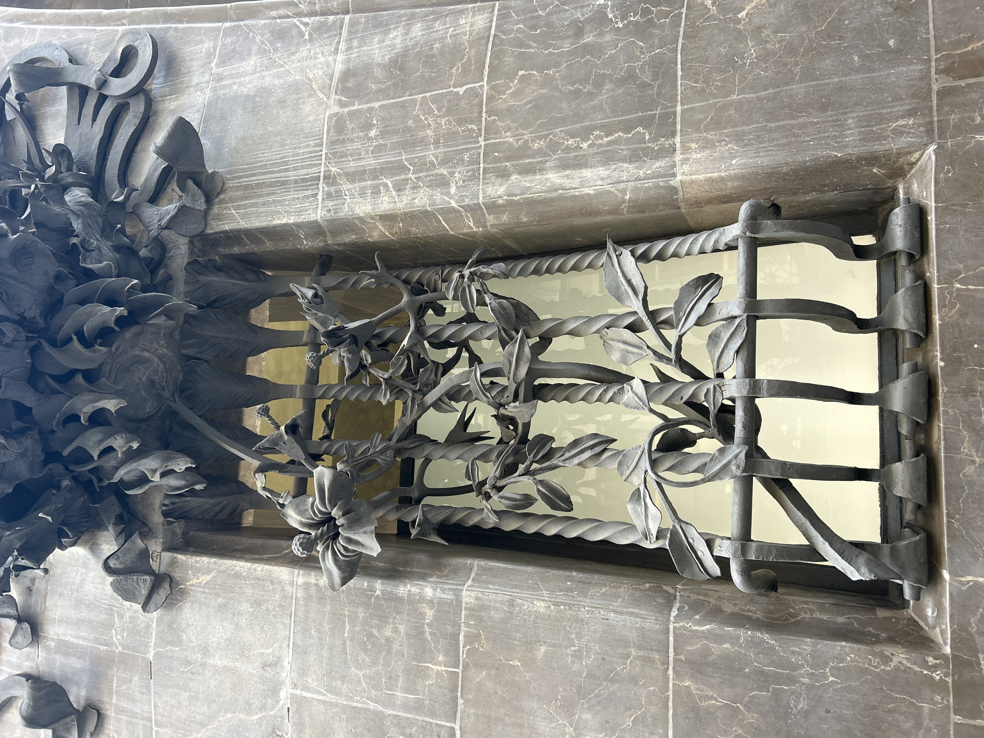 The image depicts a close-up view of ornate ironwork on a building facade in Barcelona, Spain. The facade is constructed of grey marble with distinctive white veining. The ironwork consists of a rectangular window grill with horizontal twisted bars and vertical flat bars. A stylized floral motif extends from the left side, cascading across the grill. The background behind the grill is a pale yellow. The image suggests architectural detail, possibly from a notable building in the city, although further contextual clues are not present to ascertain the specific location.