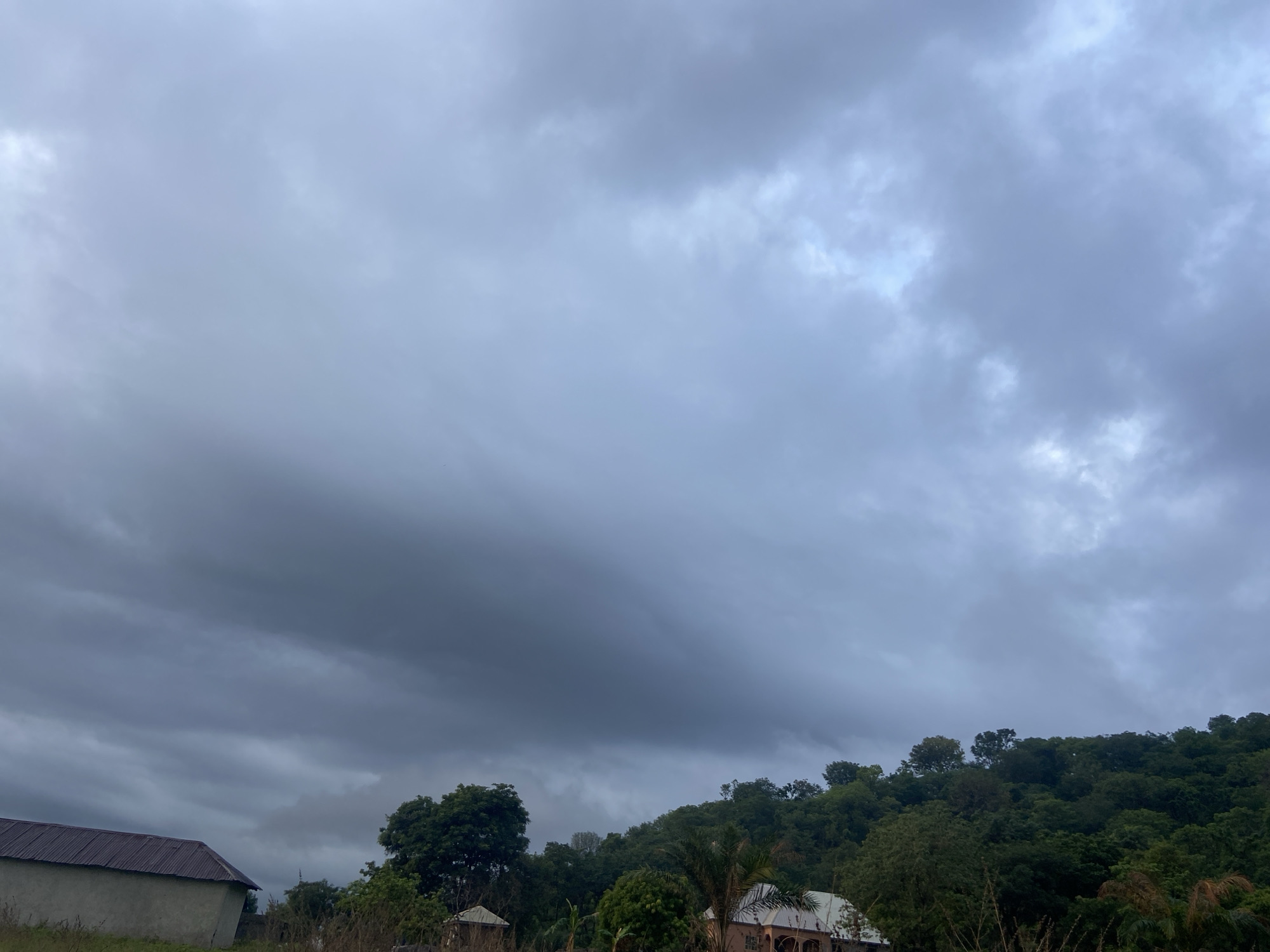 The image depicts an overcast sky over a landscape in Mararaba, Nigeria. The sky is filled with layers of gray and dark clouds, hinting at possible rain. Below the clouds, a green hillside covered with trees rises in the distance. In the foreground, there are several buildings, including a structure with a corrugated metal roof on the left, and another building with a lighter-colored roof near the center. There are also some trees and vegetation, including banana trees, scattered around the buildings. The overall scene suggests a rural or semi-rural environment under a cloudy sky.