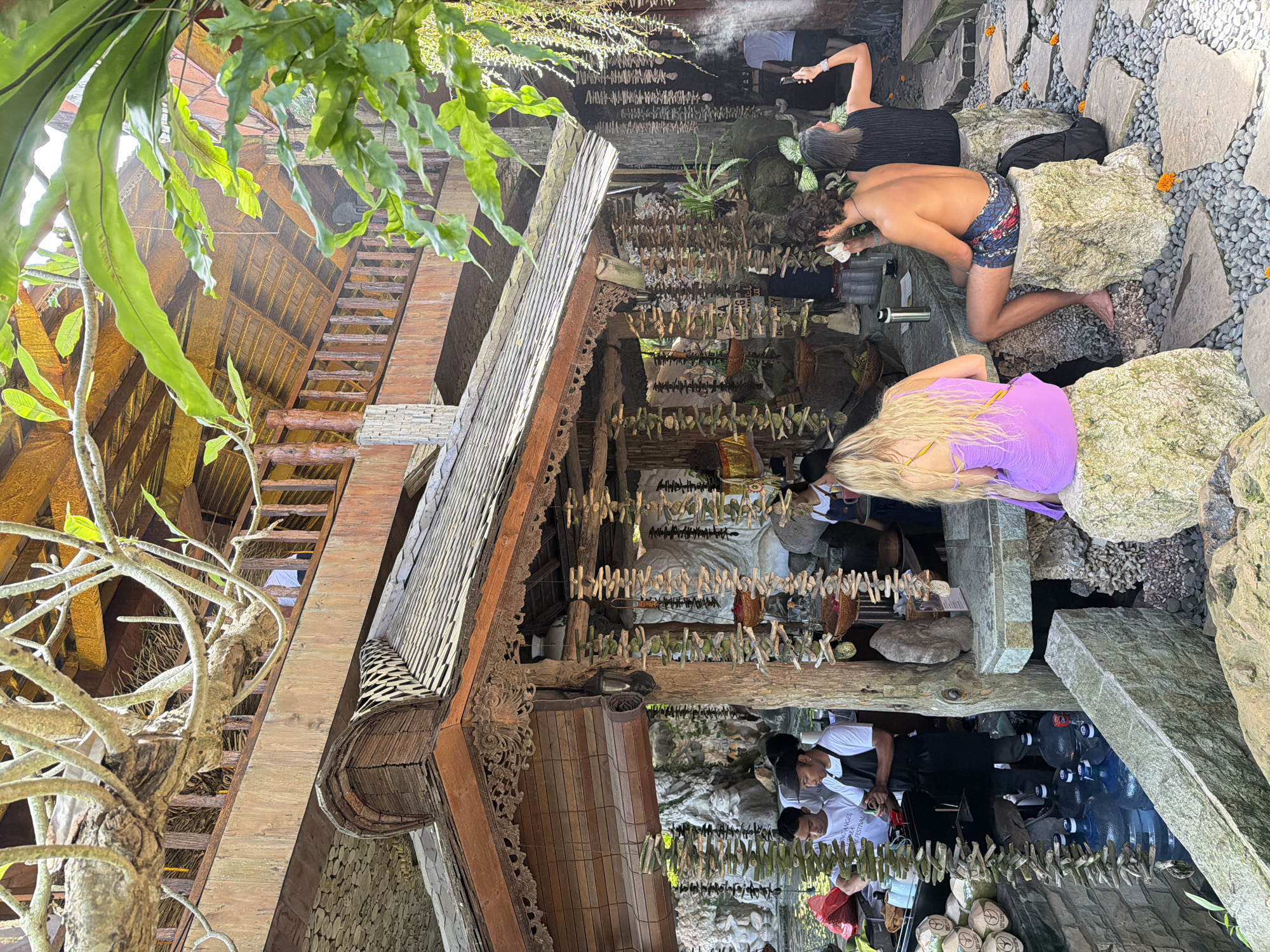 In Mengwi, Indonesia, a covered outdoor seating area features a rustic, multi-level wooden structure with thatched roofing and intricate carvings. The foreground is dominated by large, rough-hewn rocks serving as seats, with a gravel path dotted with small orange flowers. A blonde woman in a purple dress is seated on a rock, facing away from the camera. To her right, a man with curly dark hair and patterned shorts is seated, holding a cup, and a woman with dark hair in a black top is standing beside him, taking a photo with her phone. Behind them, a counter is visible, with water bottles and various decorative items. In the mid-ground, several individuals, possibly staff, are working behind a counter with coconuts and water dispensers. The overall ambiance is natural and relaxed, with lush green foliage hanging from above.