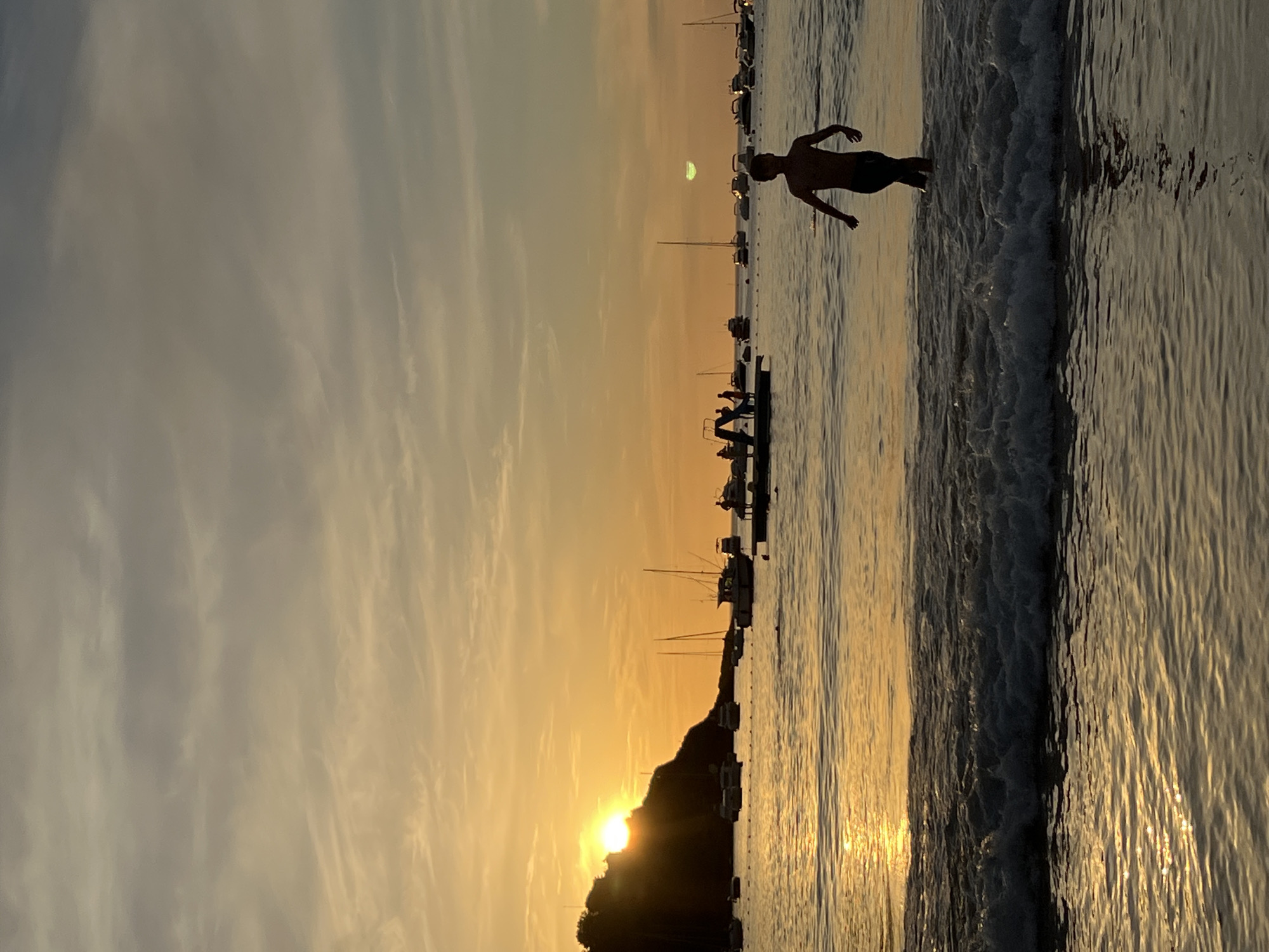A sunset scene unfolds over the Bay of La Concha in San Sebastián, Spain. The sun, partially obscured by a darkened hillside to the left, casts a warm, golden glow across the water, reflecting off the gentle waves. Several boats and yachts are anchored in the bay, their silhouettes adding to the tranquil atmosphere. In the foreground, a young person, silhouetted against the bright sky, walks out of the water. Further out, a floating platform is visible with several people on it, some appearing to be interacting. The sky above is a soft gradient of orange and grey clouds, creating a serene and picturesque setting.
