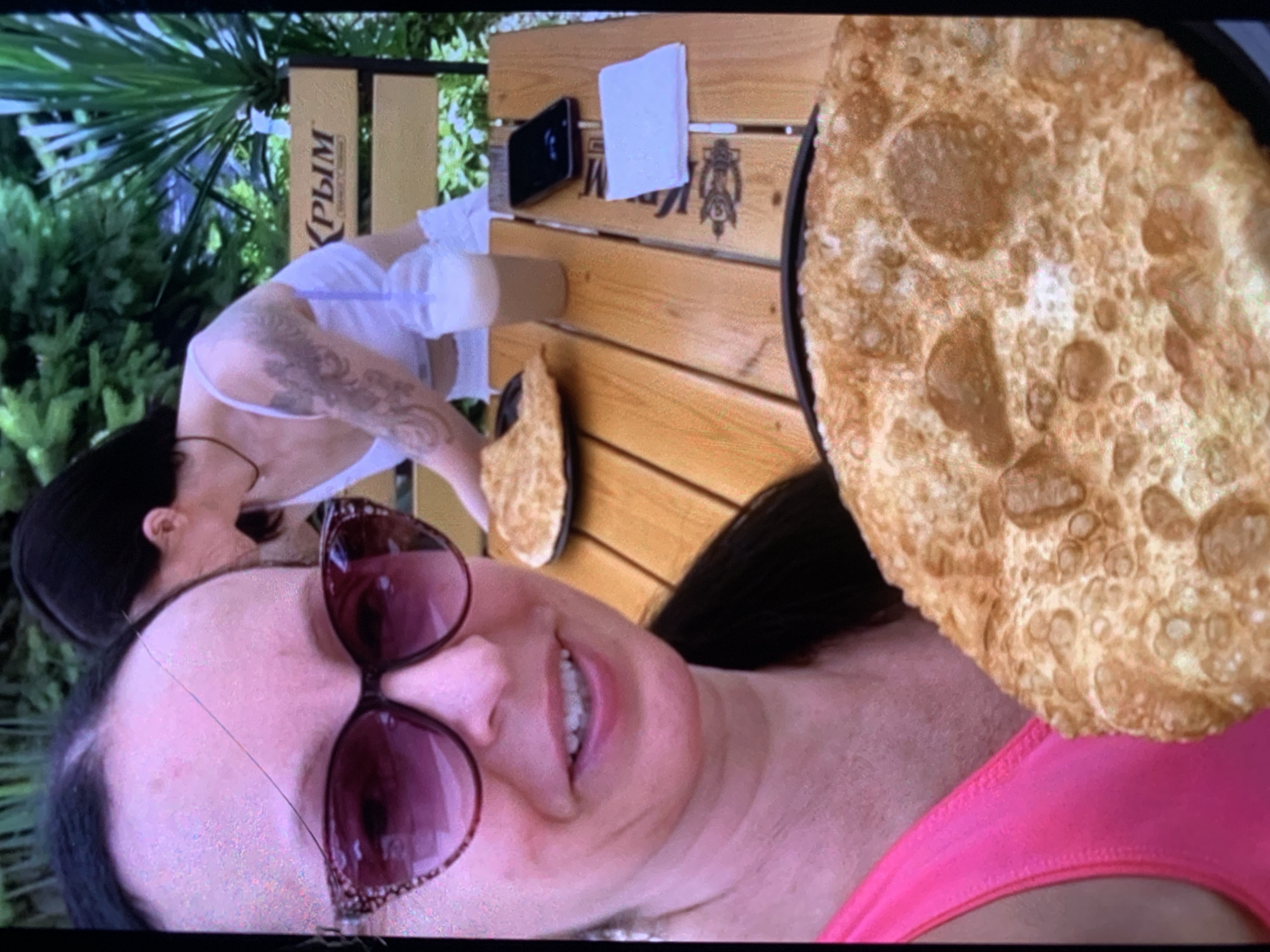 The image captures a close-up selfie taken by a woman with sunglasses and a pink top. In the background, another woman with a tattoo on her arm is visible. They are seated at a wooden table with food, drinks, and a smartphone on it. A bench with the word "Крым" (Crimea) is also in the background. The setting appears to be an outdoor cafe or restaurant.