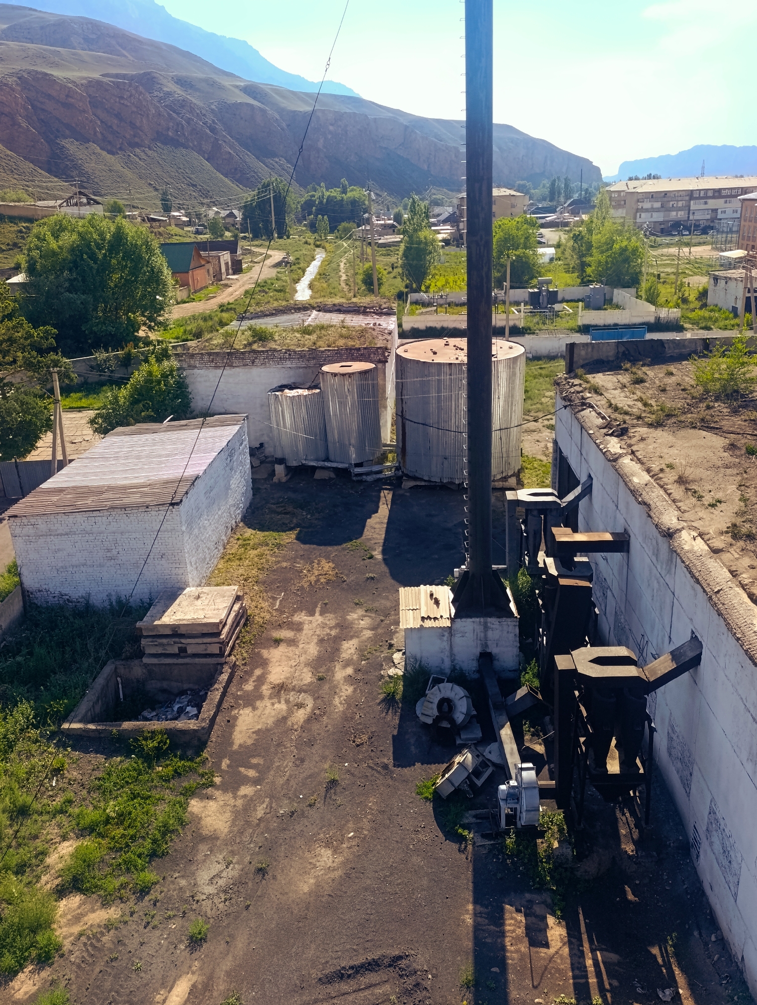 The aerial photograph shows an industrial area in Naryn City, Kyrgyzstan. In the foreground, a tall wooden utility pole dominates the center of the frame, casting long shadows. To the left of the pole is a white brick building with a corrugated metal roof, and a pile of concrete slabs. Beyond that, several large, cylindrical metal tanks are visible. To the right of the pole, a white building with a rough, discolored roof features a dark, complex industrial mechanism mounted on its exterior wall. The ground is a mixture of dirt and sparse vegetation. In the background, a stream winds through a green valley, with residential buildings and trees lining its banks. Further back, rolling hills covered in vegetation rise up, leading to a mountain range under a clear blue sky. The overall scene suggests an industrial facility situated within a rural, mountainous landscape.