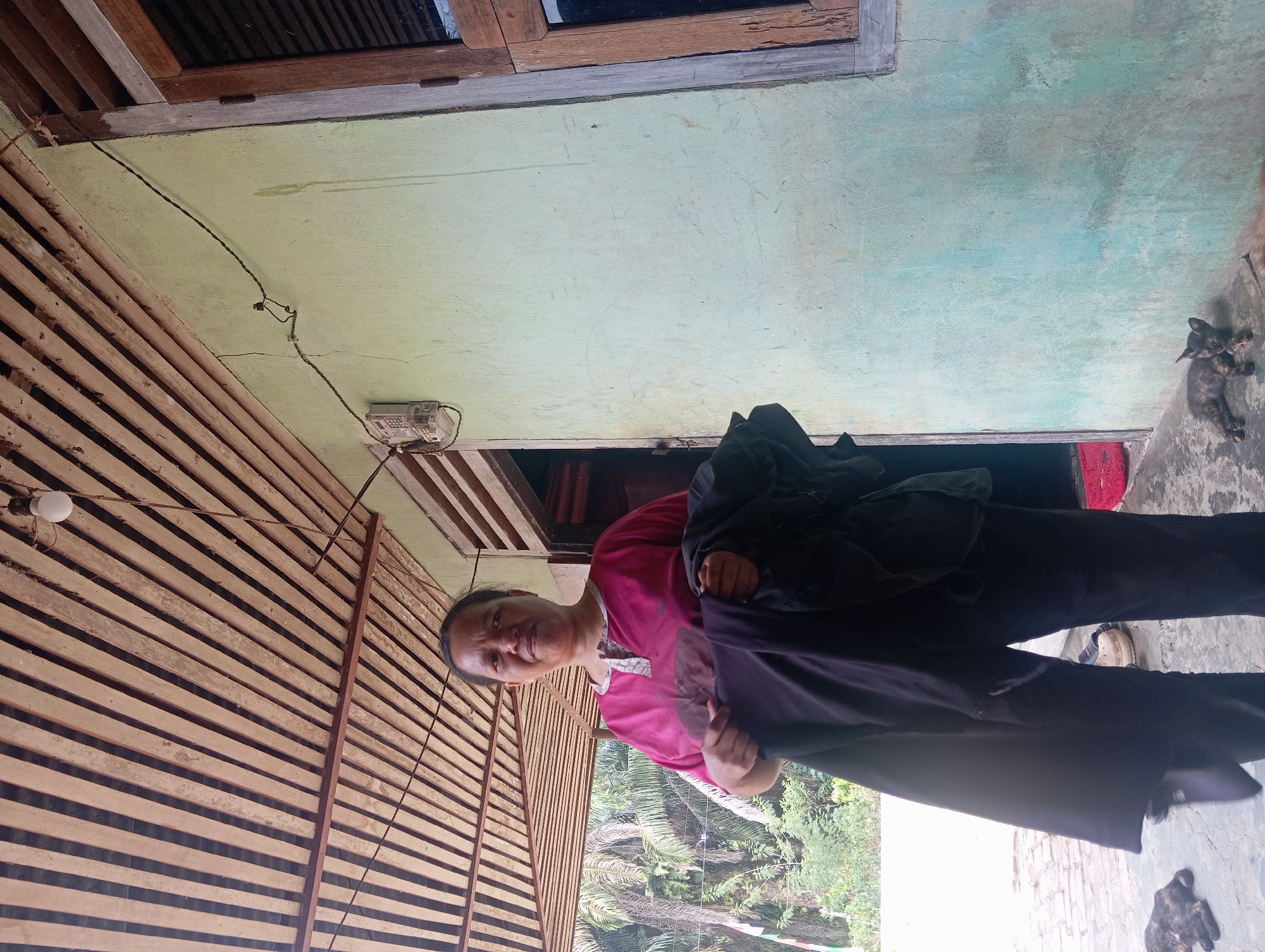 In Rejosari, Indonesia, a woman in a pink shirt stands outside a pale green house, holding dark clothing. The house has a wooden-slatted roof and a window with a wooden frame. Two cats, one dark and one lighter, are visible on the ground near the house. Trees and foliage can be seen in the background.