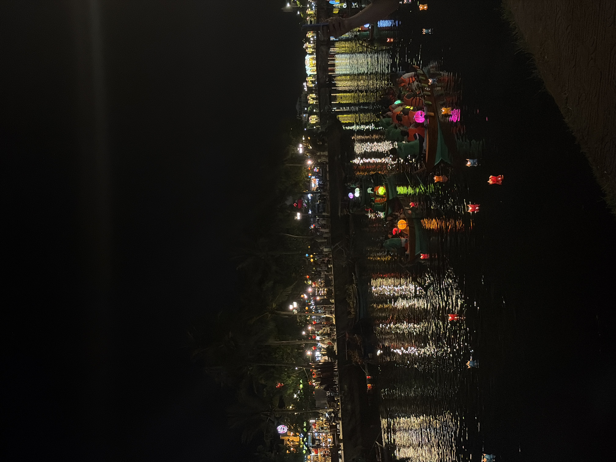 This is a vibrant nighttime photograph of a river or canal in Hoi An, Vietnam. The scene is illuminated by numerous artificial lights, creating a festive atmosphere.
In the foreground and midground, the dark water of the river is filled with reflections of lights from the riverbank and many small, colorful lanterns floating on the surface. Several traditional wooden boats are visible, carrying people, some of whom are wearing bright orange life jackets. These individuals appear to be participating in the activity of releasing or observing floating lanterns, which glow in shades of pink, red, orange, and blue.
The riverbank on the far side is brightly lit, showcasing buildings, market stalls, and silhouetted palm trees. The entire bank is adorned with a multitude of lanterns and string lights, casting warm yellow, white, and multi-colored glows that shimmer on the water. The sky above is completely dark, making the artificial illumination the dominant feature of the image. The overall impression is one of a lively and enchanting evening, characteristic of a night market or festival on the water. No discernible text is visible.