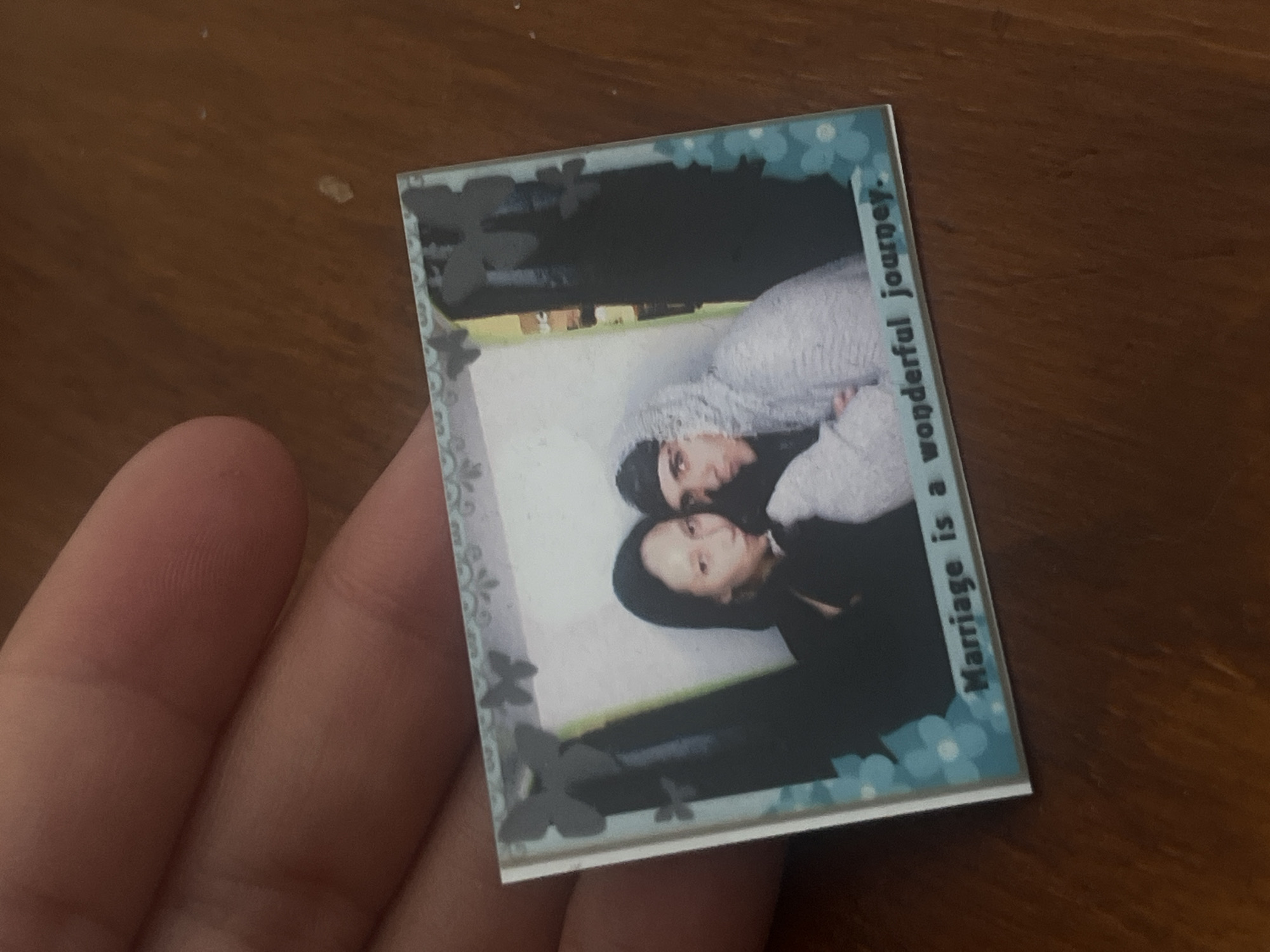 The image shows a small, framed photo being held in someone's hand. The photo features a couple, likely a man and a woman, embracing closely. The frame around the photo has a light blue floral design and the words "Marriage is a wonderful journey" printed vertically along one edge. The background of the photo within the frame appears to be outdoors, with some greenery and a building visible. There is no indication of the location being in Buenos Aires, Argentina, or any other city for that matter.