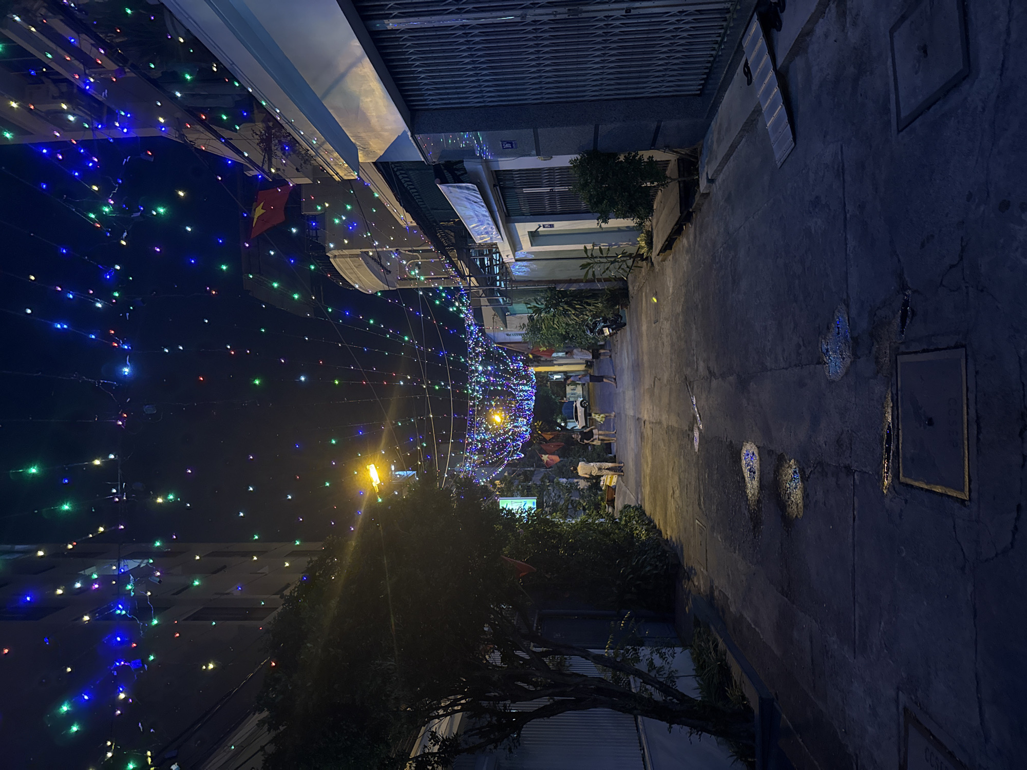 Captured at night, the image shows a narrow street in Nam Nha Trang, Vietnam, adorned with festive lighting. Multi-colored lights are strung across the street, creating an illuminated tunnel effect. A Vietnamese flag is visible among the lights. People are walking along the street, casting shadows in the dim light. The street is flanked by buildings with closed doors and parked motorbikes, typical of Southeast Asian urban scenes. The pavement is cracked and uneven, with drainage covers visible. The photograph is taken from a low angle, looking slightly upwards along the street.