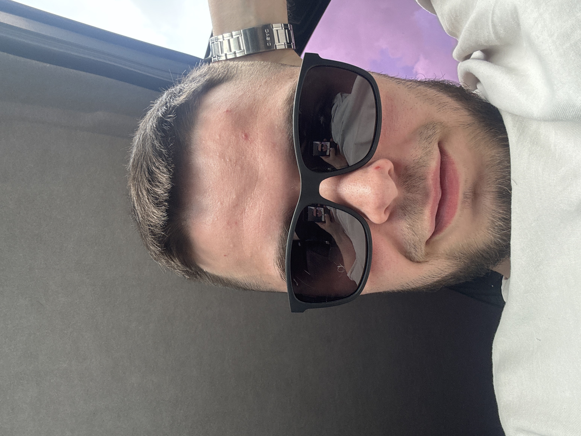 The image is an upward-angled selfie of a person wearing sunglasses and a white hoodie. The person is a man with short, light brown hair and stubble. He has a silver watch on his left wrist with the brand "CASIO" visible. The background includes a purple sky with clouds, suggesting it's either daytime with colorful clouds or dusk/dawn. Part of the setting appears to be indoors, as indicated by a grey wall or surface behind the person. The lighting suggests it might be a bright day, possibly outdoors or near a window. The person's expression is neutral, with a slight smile. There are no clear indicators of a specific event taking place. The image seems to be taken in Perm, Russia.